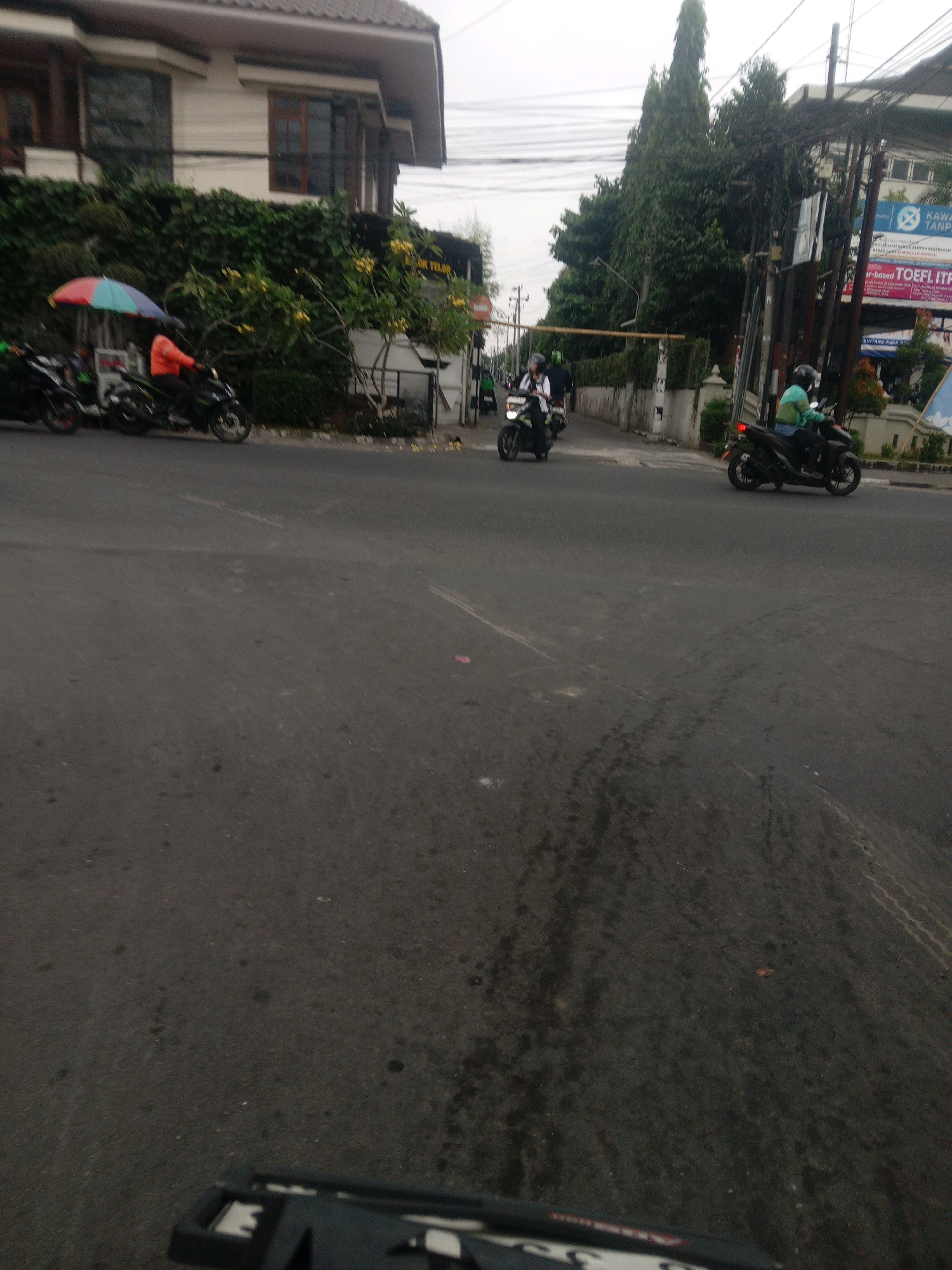 This is a low-angle, horizontal shot of a street scene in Depok, Indonesia, likely taken from a moving vehicle. In the foreground, a dark asphalt road displays tire marks. The midground features several motorcycles. One motorcycle, with a rider in an orange jacket, is parked under a colorful umbrella. To its right, another motorcycle is moving away. Further down the road, more motorcycles are visible, some with riders. The background includes a building with a white facade and brown window frames, overgrown green foliage, and a line of tall trees. Electrical wires and poles are prominent, along with street signs and a building with a blue sign that appears to advertise TOEFL preparation. The sky is overcast.