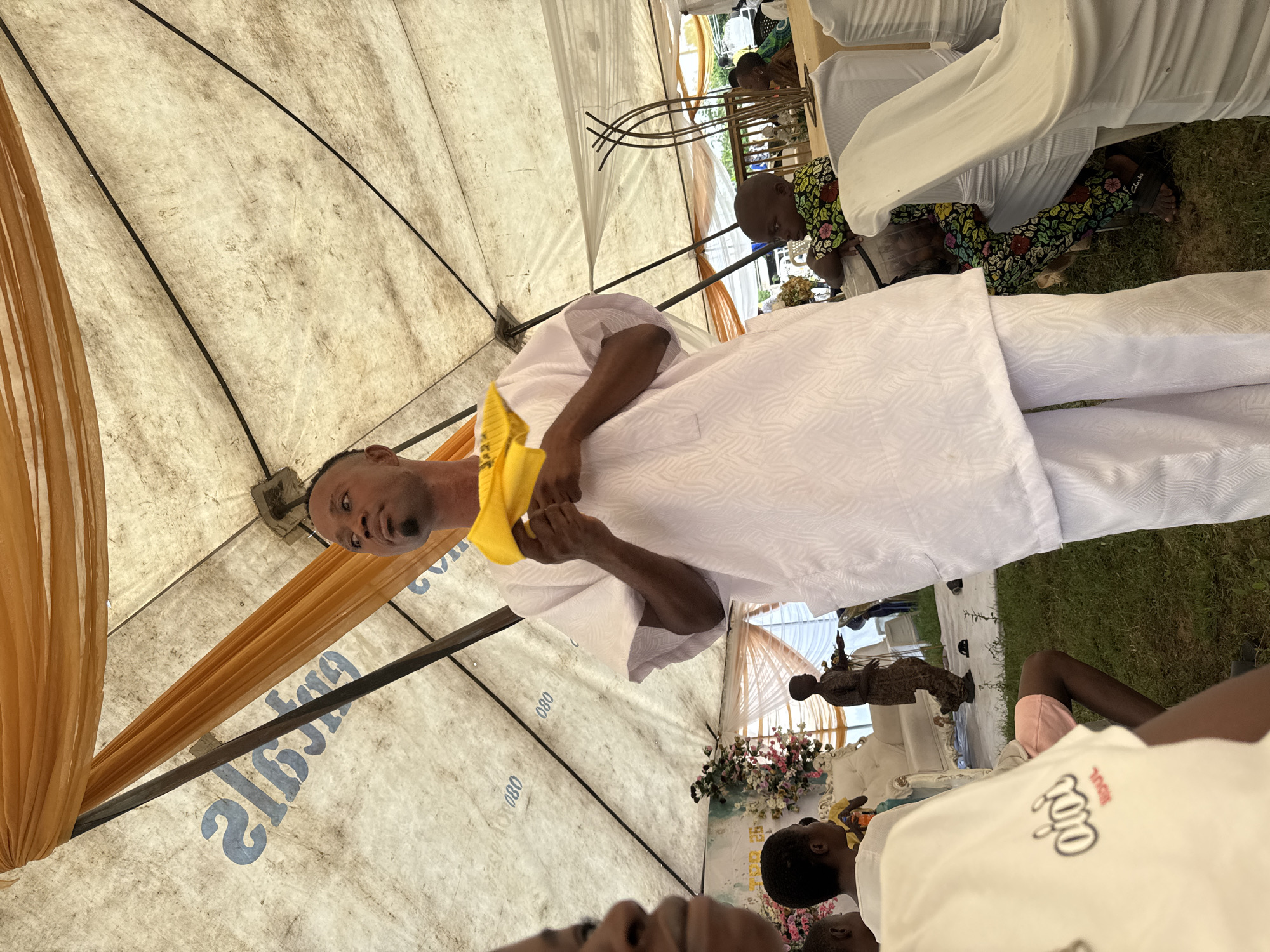 The image depicts an outdoor gathering under a large white tent with orange fabric draped from the poles. In the foreground, a man dressed in white traditional attire, holding a yellow object, is speaking. He has a goatee and short hair. To the left of the man, a child's face is visible, also in the foreground.
In the background, several other people are present. A child in a floral patterned outfit is seated at a table. Another child, dressed in traditional brown clothing, is standing near a white sofa adorned with flowers. The ground is covered with green grass. The tent's interior features black poles and visible fabric seams. The tent fabric also has blue lettering that reads "080" multiple times.
This scene is likely from a social event or ceremony taking place in the city of Badagry, Nigeria, given the cultural attire and the style of the tent setup often seen at such gatherings.