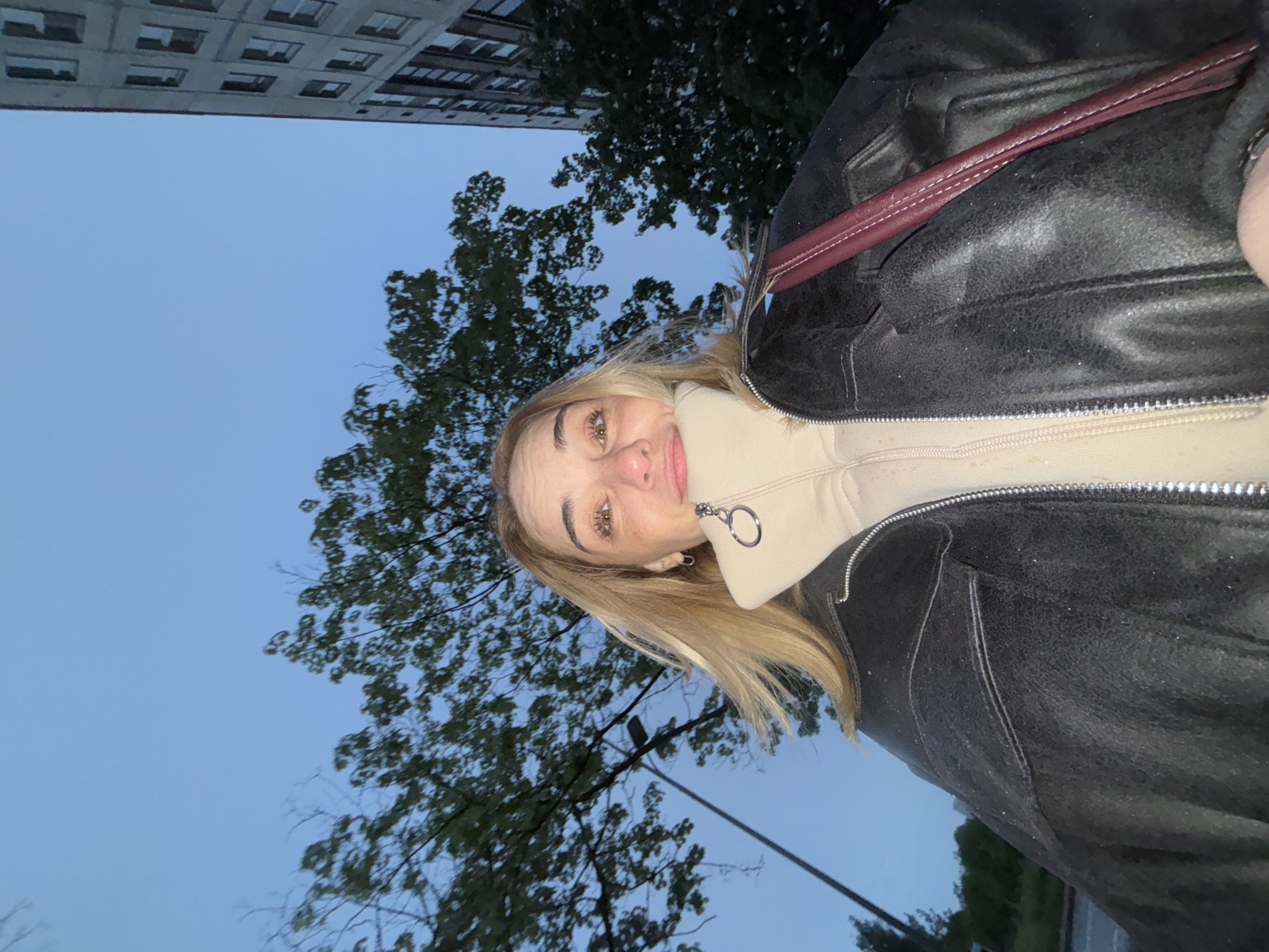 A woman with blonde hair is taking a selfie outdoors. She is wearing a black leather jacket over a light-colored zip-up hoodie. A maroon strap is visible on her jacket. In the background are trees, a building, and a blue sky.
