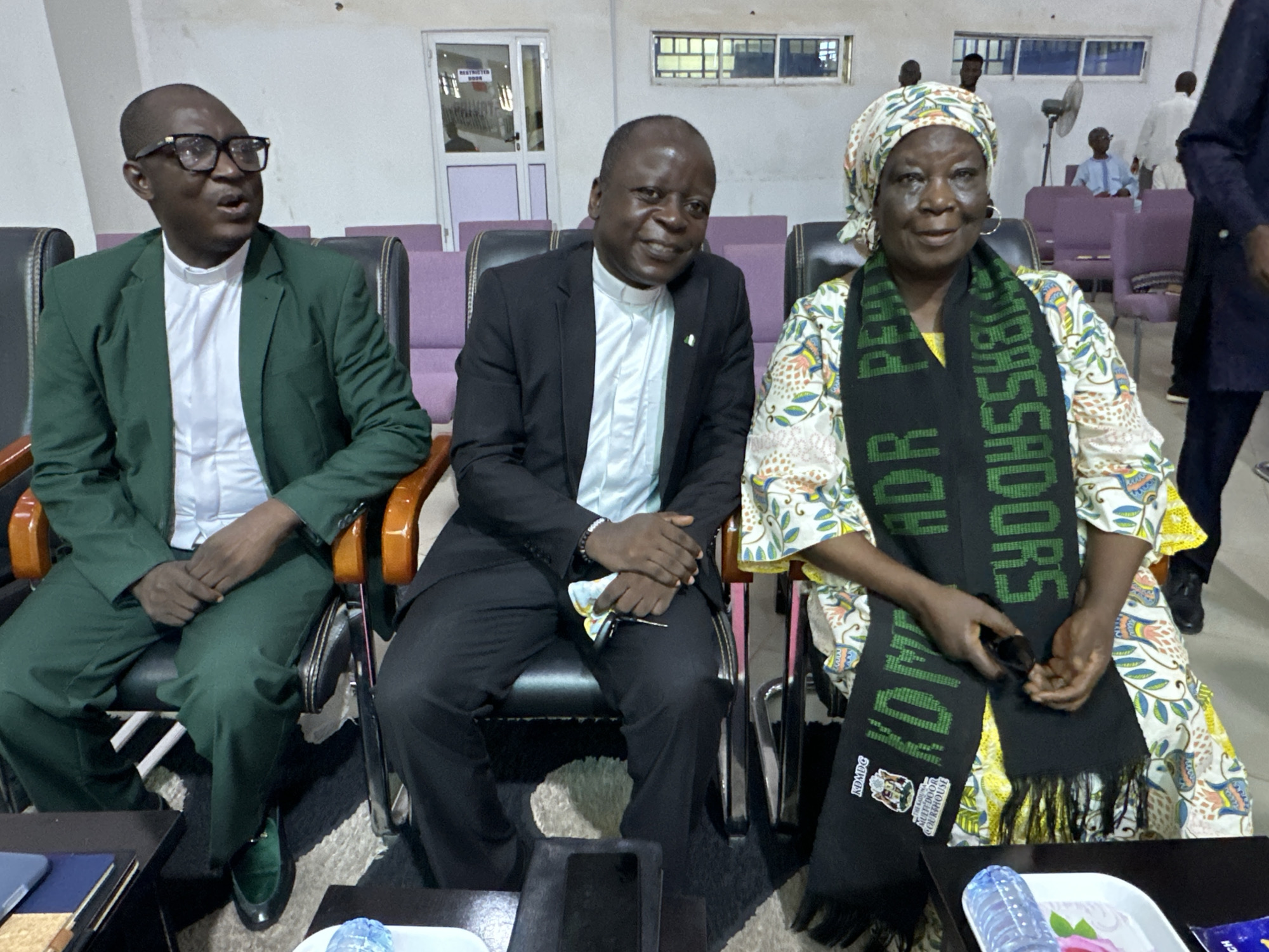 In Goni Gara, Nigeria, three individuals are seated in a room that appears to be an event venue or meeting space. The room has rows of chairs, some purple and some black with wooden accents, and the walls are a neutral color. In the background, there is a door with "RESTRICTED ACCESS" signage and windows.
On the left, a man wearing glasses and a green suit over a white clerical collar is seated. To his right, a man in a black suit and white clerical collar is smiling. On the far right, a woman wearing a colorful patterned dress and headscarf is seated, with a black scarf around her neck that has green text. She is holding her hands in her lap. The people are seated at tables, and a bottle of water is visible on a white tray in the foreground. The overall atmosphere appears to be formal or ceremonial.
