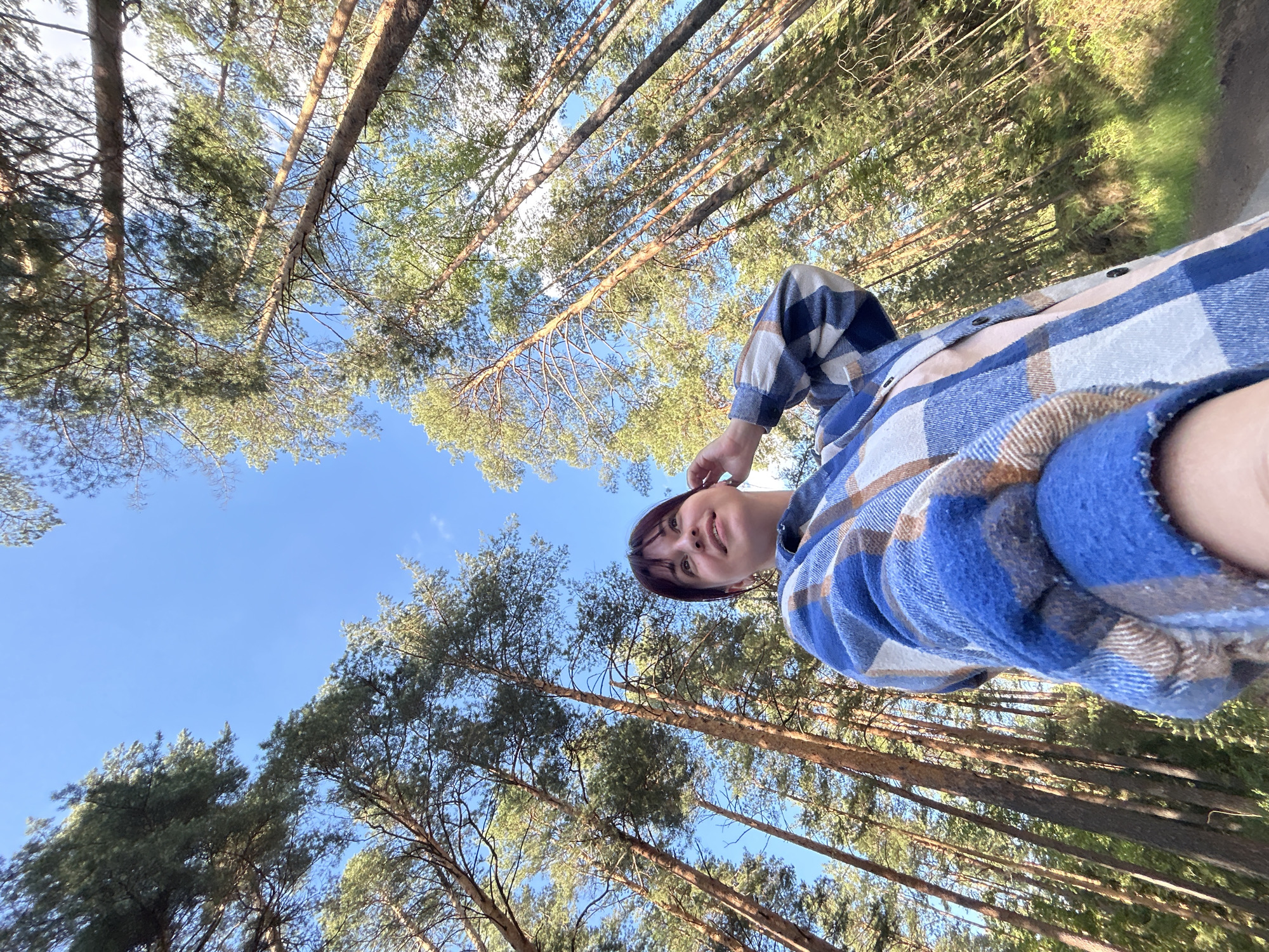 A young woman with dark hair and bangs is captured in a low-angle selfie, standing in a forest of tall pine trees. She is wearing a blue, white, and brown plaid shirt. The woman is looking up and to her right with a slight smile, her right hand touching her hair. The background is dominated by the clear blue sky and the towering, slender trunks of pine trees, with their green needles forming a dense canopy. A dirt path is visible in the lower right corner of the image. The lighting suggests it is daytime, possibly late afternoon due to the warm tones. The location is identified as the city of Fokino, Russia.