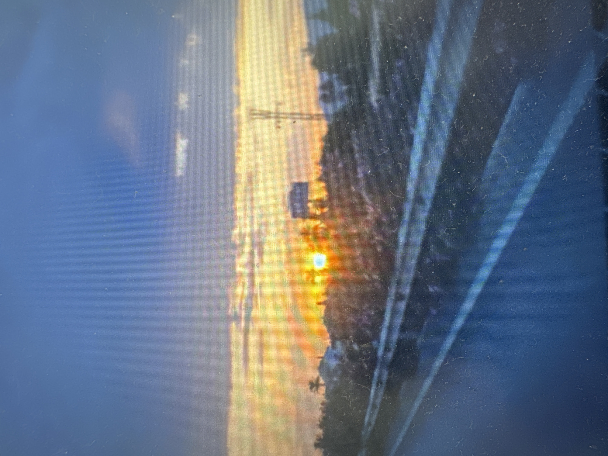The image depicts a sunset over a landscape that appears to be on the outskirts of Murcia, Spain, characterized by a highway in the foreground. The sun, a bright orb, is low on the horizon, casting a warm glow across the sky and illuminating scattered clouds with hues of orange and yellow. Silhouetted against the vibrant sky are trees, some palm trees, and what appears to be a building, possibly a villa or a residential structure. A prominent feature in the mid-ground is a large billboard or sign, and further in the distance, a power line tower stands tall. The foreground shows a highway with multiple lanes, delineated by white lines, suggesting a transportation artery. The overall impression is one of a tranquil, late afternoon or early evening scene, with the natural beauty of the sunset contrasting with the man-made elements of the highway and infrastructure. The image appears to be captured from a moving vehicle, given the perspective and slight blur.