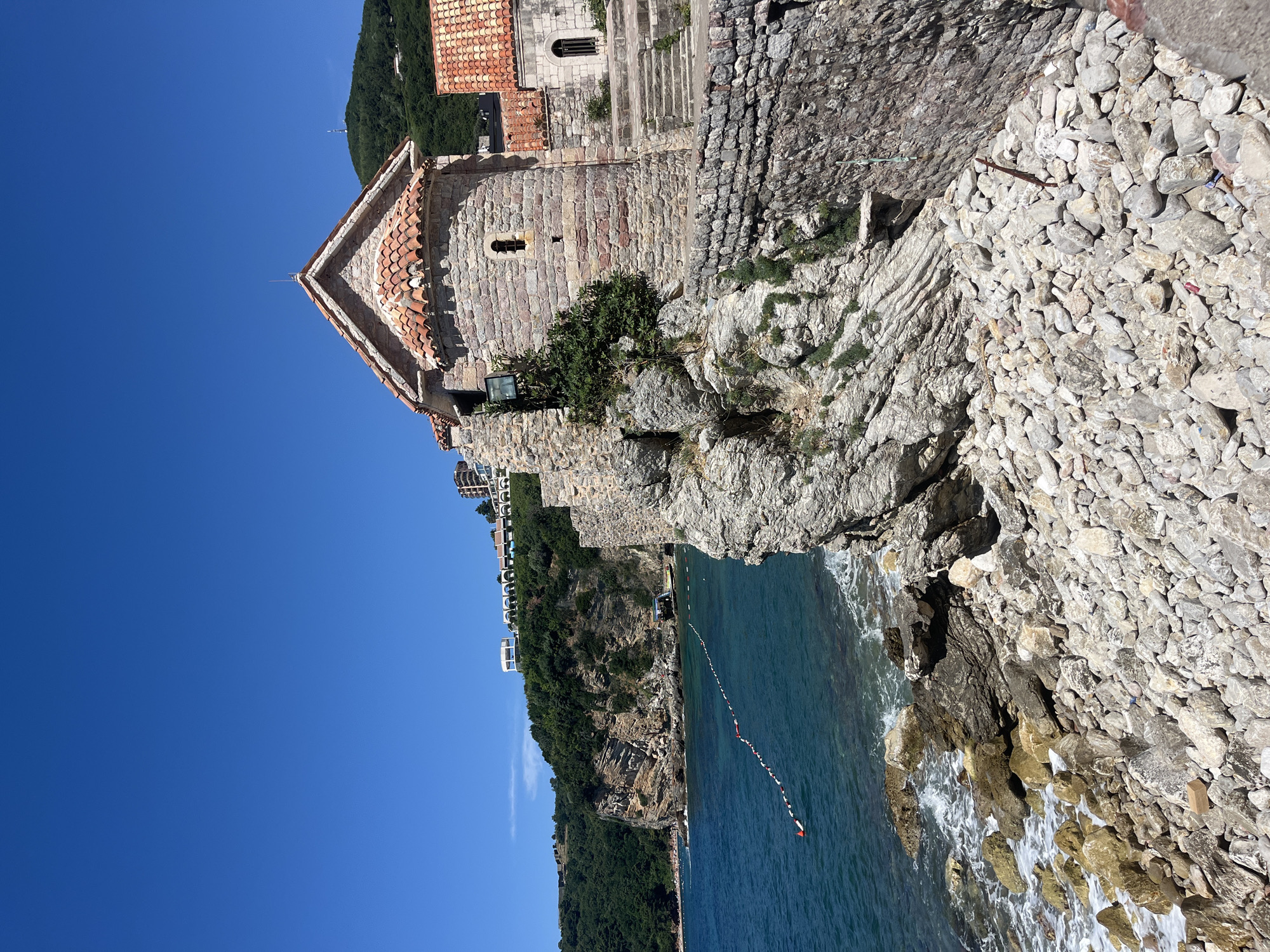 The image depicts a sunny day scene in Budva, Montenegro, focusing on a stone building situated on a rocky cliff overlooking the Adriatic Sea. The building, with a terracotta tiled roof and small arched windows, appears to be an old church or a historical structure. Below the building, the sea is a vibrant blue, with gentle waves lapping against the rocky shore. A line of buoys with red and white markers stretches across a section of the water, possibly indicating a swimming area.
In the background, lush green hills rise from the coastline, dotted with buildings and a white structure with arched windows near the summit. The terrain is rugged, with visible layers of rock formations on the cliff face. The foreground is dominated by a dense scattering of grey and white stones and rocks, leading down to the water's edge. The sky is clear and bright blue, suggesting fair weather. There are no people visible in the immediate foreground or on the structure, but the buoys in the water suggest human activity in the vicinity. The scene evokes a sense of historical charm and natural coastal beauty.