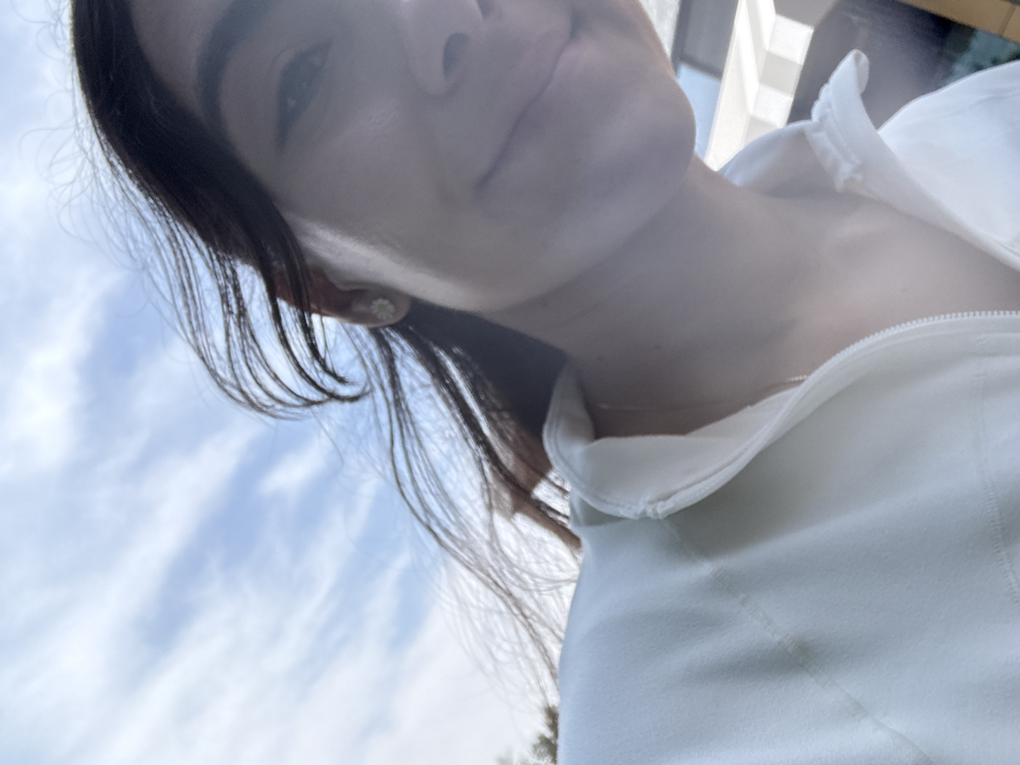This image, taken in Puebla, Mexico, captures a close-up, upward-angled view of a person, likely a female, from the chest upwards. Her head is tilted slightly down, and she appears to be looking towards the camera with a serene, neutral expression, possibly with a subtle smile. She has dark hair that frames her face and flows downwards on the left side of the frame. A small, white daisy-shaped stud earring is visible on her left ear. She is wearing a light-colored, possibly white or off-white, collared top with a visible zipper closure down the front.
The background primarily features a bright, light blue sky with scattered wispy white clouds, indicating an outdoor setting during a clear day. The lighting is soft and natural, illuminating the subject gently. Out-of-focus elements, potentially distant structures or foliage, are subtly visible in the upper right portion of the image. There is no visible text in the media file. The overall atmosphere conveyed is calm and natural.