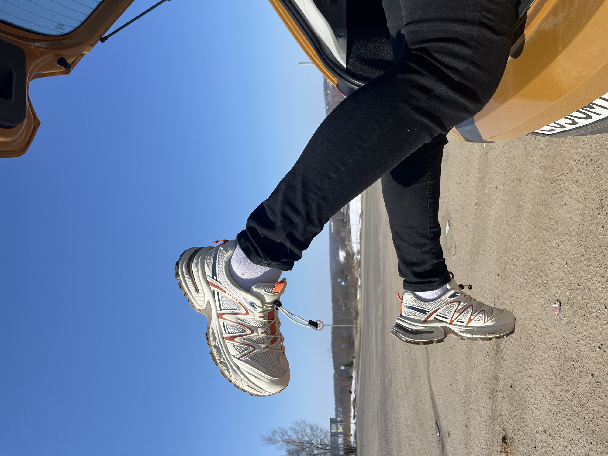 The image captures a close-up view of two legs, clothed in black pants and white socks, extending from the open trunk of a mustard-yellow vehicle. The feet are fitted with white, grey, and orange athletic sneakers with prominent soles. The car is situated on a light brown, unpaved surface. A partial Russian license plate, displaying characters such as 'У050УМ', is visible on the lower rear of the vehicle. Beyond the vehicle and immediate foreground, a paved road or intersection is visible. The background reveals a rural landscape under a clear blue sky, featuring bare deciduous trees, distant hills, and patches of snow, along with a circular red-bordered road sign. The scene is an outdoor setting in Zubovo, Russia, observed under bright daylight.