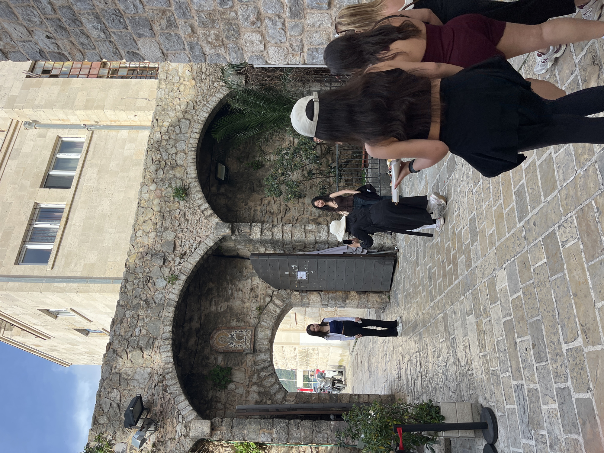 The image shows a cobblestone street scene in Budva, Montenegro, featuring a stone archway with two upper arches framing views further down the passage. At the front of the image, several individuals stand or lean, occupying the upper-right corner. Further into the scene, a person in a black dress and hat seems to be taking a photo with a smartphone. Deeper into the archway, a single person in a light top and dark pants stands near the end of the passage. The architecture is primarily stone, with a building visible on the left side with multiple windows. Light levels decrease as the passage extends into the distance, where hints of urban activity, possibly traffic, are visible.