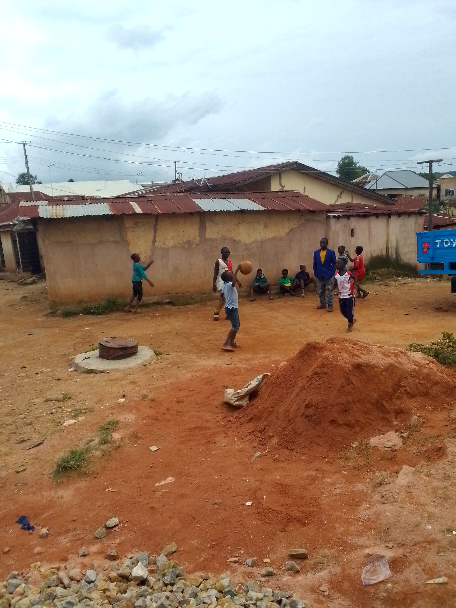 The image depicts an outdoor scene in a dusty, reddish-brown dirt area, possibly a residential or informal settlement. Several young boys are engaged in a game of soccer. One boy in a blue shirt and black shorts is mid-kick, another in a white and red jersey with dark pants is running, and a third boy in a light blue shirt and dark shorts is reaching out, possibly to catch or pass the ball. To the left, a boy in a green shirt and dark shorts is kicking a ball upwards, while other boys sit and watch. In the background, a tan-colored building with a corrugated metal roof stands, and further back, a blue truck with "TOYO" written on it is parked. The sky is overcast. The ground is uneven and scattered with small stones and patches of dry grass. A concrete manhole cover is visible in the foreground. A pile of red dirt is prominent in the lower right quadrant.