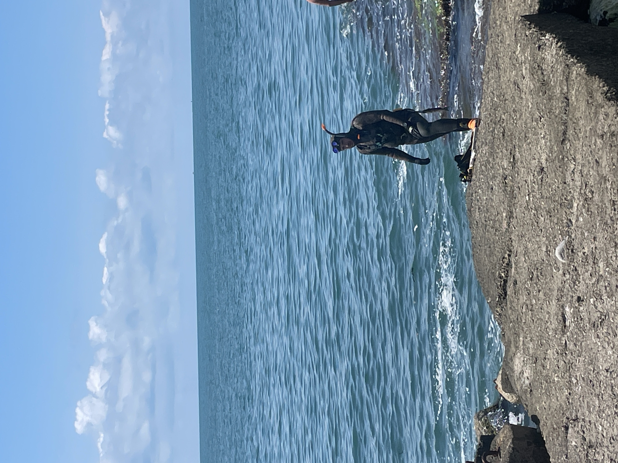 In Sochi, Russia, a person dressed in a black wetsuit, snorkel, and goggles stands on a concrete breakwater. The breakwater is rough and uneven, with some rocks and debris scattered on its surface. To the right of the breakwater, the ocean is visible, with waves gently lapping at the shore. The water is a deep blue, and the sky above is a clear, bright blue with a few fluffy white clouds. The scene appears to be a sunny day at the beach, with the person preparing to enter the water.