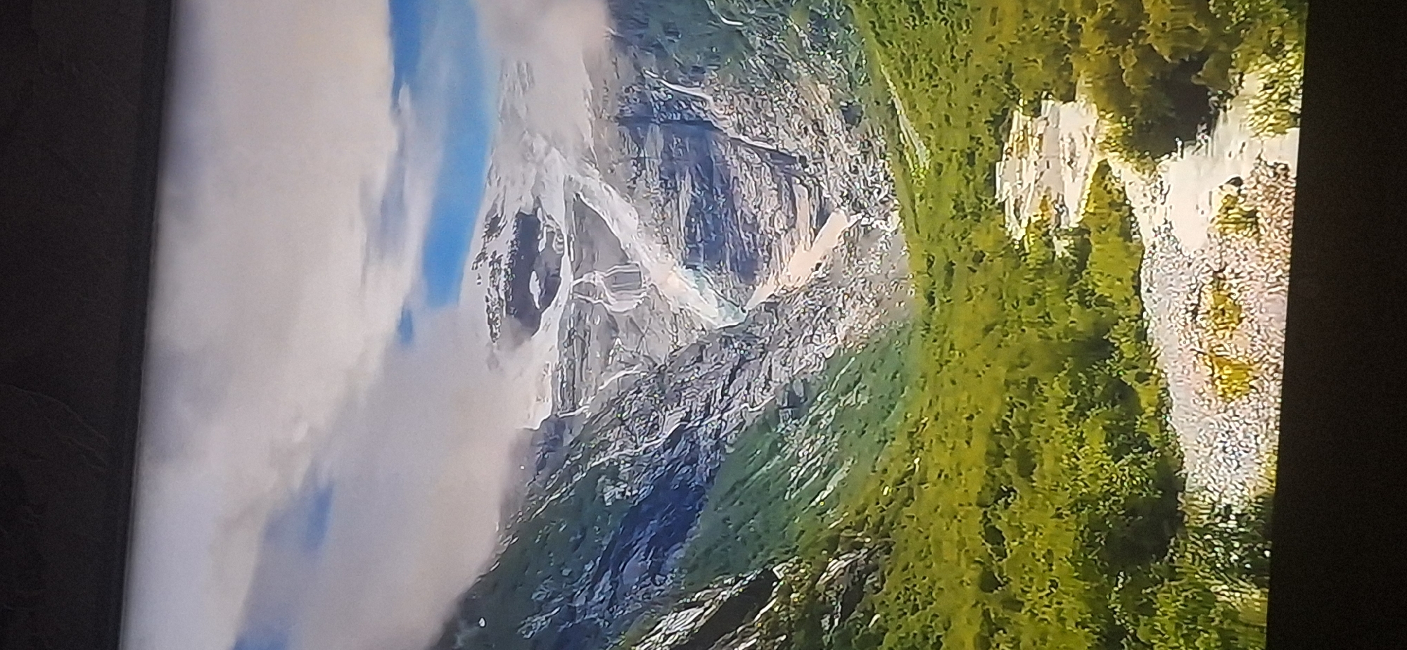 The image depicts a majestic mountain landscape featuring a glacier at the summit. A river flows through a lush green valley, winding its way towards the foreground. The sky is partially cloudy with patches of blue visible. There are no people or specific urban features visible in this natural scene. The location of Bila Tserkva, Ukraine is not depicted in this image.