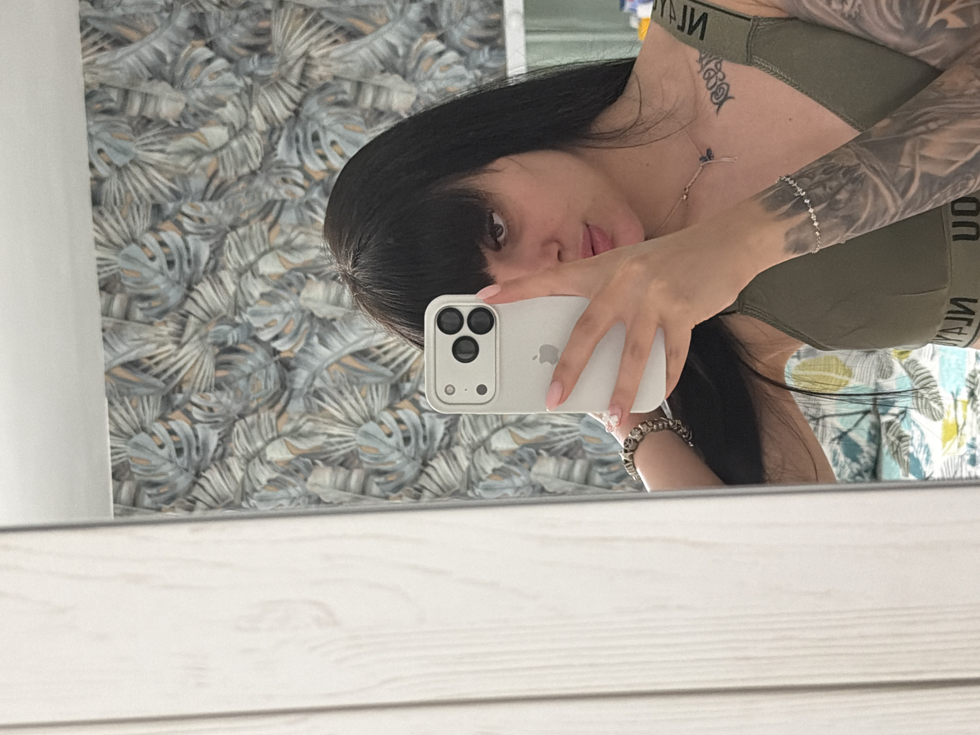 A person takes a mirror selfie in an indoor setting. The individual has long, dark hair with bangs and is wearing an olive green sports bra or crop top, with visible mirrored text on the elastic band that appears to read "УЛJИ" or "УГИ". Their face is partially obscured by a light-colored iPhone, held in their left hand, which features three camera lenses in a triangular configuration and an Apple logo on its back. The person has a full sleeve tattoo on their right arm and a smaller dark tattoo on their upper left chest/shoulder area. They wear a thin, silver-toned bracelet on their left wrist and a thicker, darker bracelet on their right. A silver-toned necklace with a small, dark blue or black pendant and a dangling silver element is also visible. The background, reflected in the mirror, displays a wall covered with patterned wallpaper featuring stylized leaves in shades of grey, light blue, and muted orange. A white ceiling is visible at the top, and a light-colored wooden structure is partially reflected on the far left. A blurred glimpse of a patterned bed or duvet is visible in the lower right portion of the reflection.