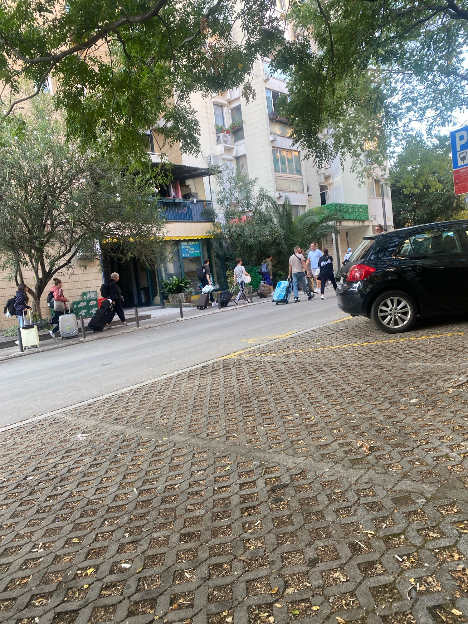 In Budva, Montenegro, a group of individuals are walking along a paved street, some pulling luggage. The scene is bordered by a building with balconies and trees, suggesting an urban environment. A black car is parked on the right side of the street. The foreground features a decorative concrete paver pattern.