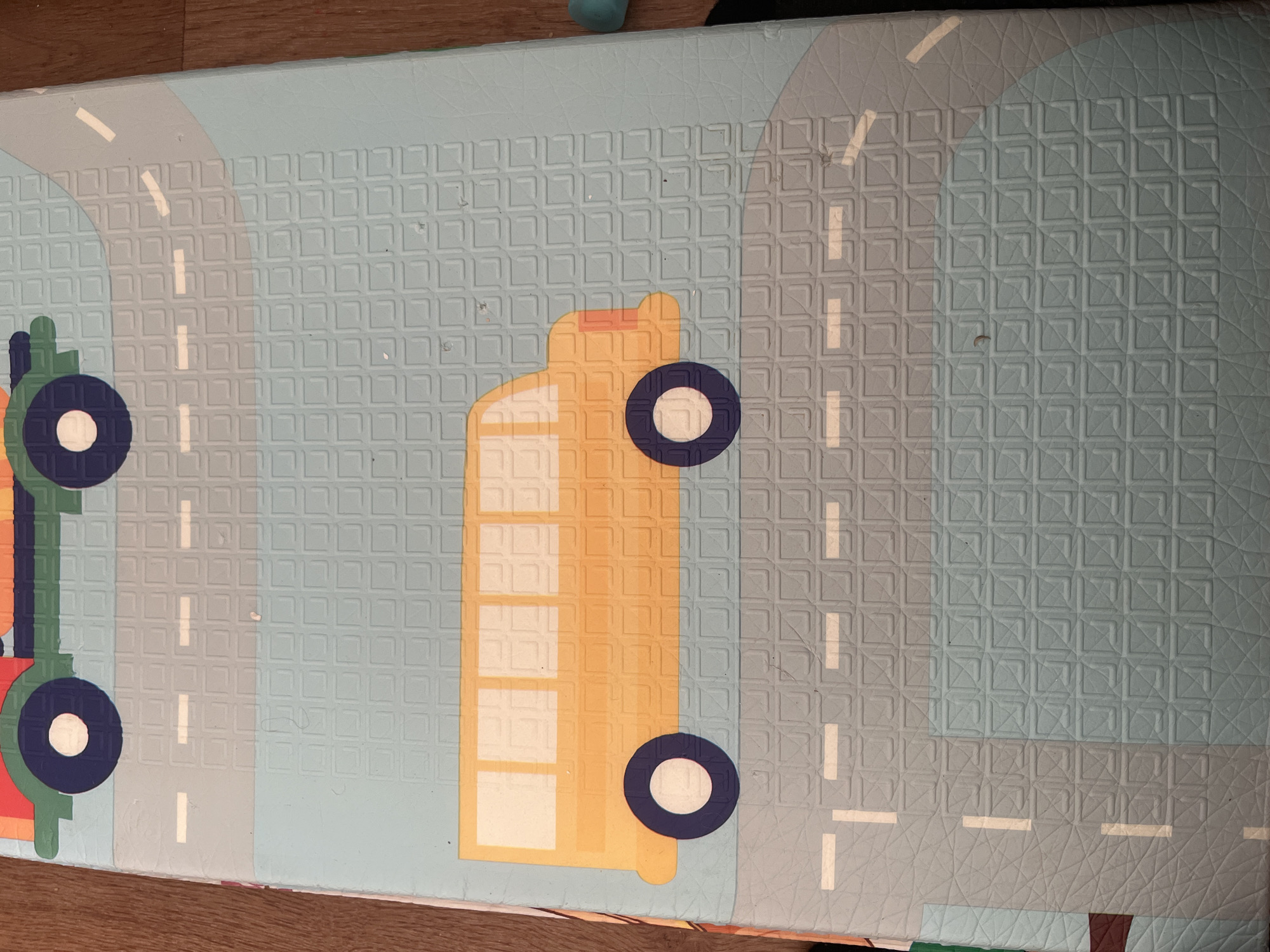 The image is an overhead, slightly angled view of a brightly colored play mat, likely for children. The mat is designed to resemble a city street scene with various vehicles and road markings. Dominating the foreground is a yellow school bus with white windows and blue wheels. In the background, a portion of another vehicle, possibly a car or truck, with green and red elements and blue wheels is visible. The mat has a textured surface with raised square patterns. The background is a light blue with gray road segments and white dashed lines indicating lanes. The lighting is even, suggesting indoor illumination. The lower right edge of the mat shows a sliver of a wooden floor. The scene is static and depicts a toy environment. There are no people or animals present. No identifiable landmarks or street signs are visible to suggest a specific location within Chelyabinsk, Russia, or any other city.