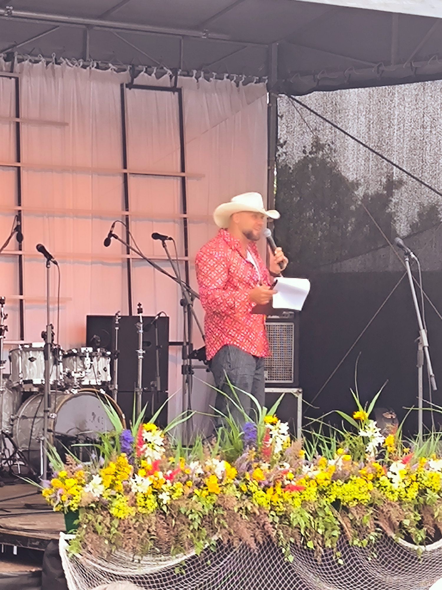 A man wearing a white cowboy hat, a red patterned shirt, and dark pants stands on a stage at the Positivus Festival in Salacgrīva, Latvia, holding a microphone and a piece of paper. He is speaking into the microphone, and his mouth is open. Behind him, a drum set, speakers, and microphones are visible. In front of the stage, a floral arrangement adorns the front of the stage. The backdrop consists of white curtains and a dark textured material. The man appears to be a performer or host.