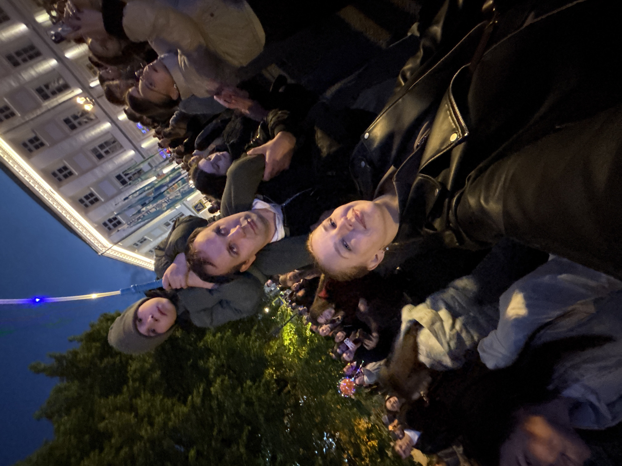A nighttime outdoor scene depicts a crowd of people, possibly at an event, with a building visible in the background. The building has multiple windows and is illuminated with lights. In the foreground, a group of individuals are gathered, some wearing casual clothing. The focus is on the people in the foreground. In the bottom left, a tree is partially visible. The specific location in Podosenikha, Russia cannot be conclusively determined from this image.