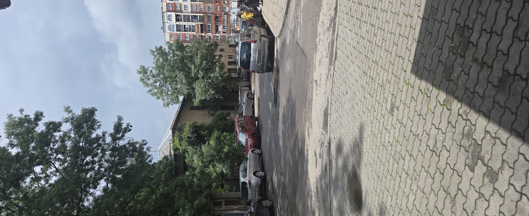 This media file captures an urban street scene on a bright, sunny day. The image is oriented vertically (or rotated 90 degrees counter-clockwise), showing a paved sidewalk with a distinctive wavy pattern on the right, an asphalt road in the center, and buildings and trees on the left.
Numerous cars are parked along the left side of the road, facing away from the viewer. A silver SUV with the license plate "B 9316 CB" is prominently featured, and another silver car further down the street has a partially visible license plate "B 4516 CB". These license plates, starting with "B", indicate the location is likely Bulgaria.
The left side of the scene is dominated by lush green trees, with their foliage extending high into the frame. Behind the trees, a multi-story residential building with a varied facade of brick and painted sections, numerous windows, and balconies is visible. Another, older-looking building with a rusty metal roofline is partially visible amidst the trees closer to the foreground.
The sky is largely clear with some scattered white clouds. Strong sunlight casts distinct, elongated shadows across both the road and the patterned sidewalk, suggesting it is daytime, possibly late morning or early afternoon. In the distance, near the tall residential building, a few indistinct human figures can be observed walking, but no specific activities are discernible beyond general street life. No explicit emotions are conveyed.