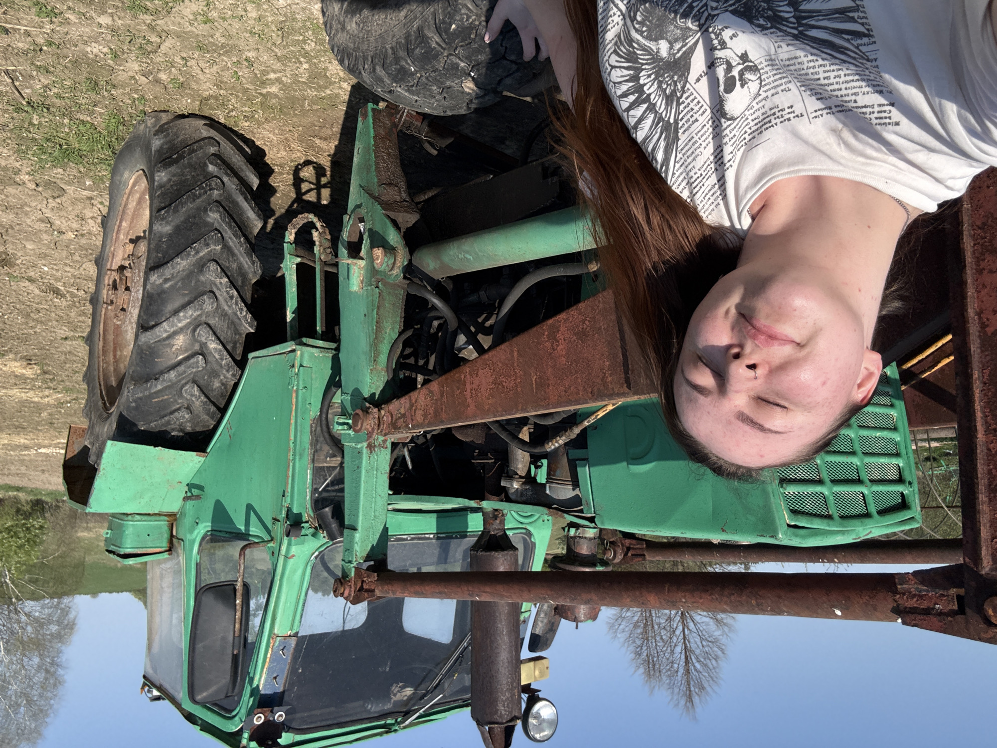 The image shows a woman lying on the front of a green tractor. The woman has brown hair, closed eyes, and a nose ring. She is wearing a white t-shirt with black print. The tractor appears old and rusty in places, with large tires and a metal frame. The background suggests a rural environment with trees and a blue sky. There is no information in the image that indicates the location as the city of Lukintsy, Russia.