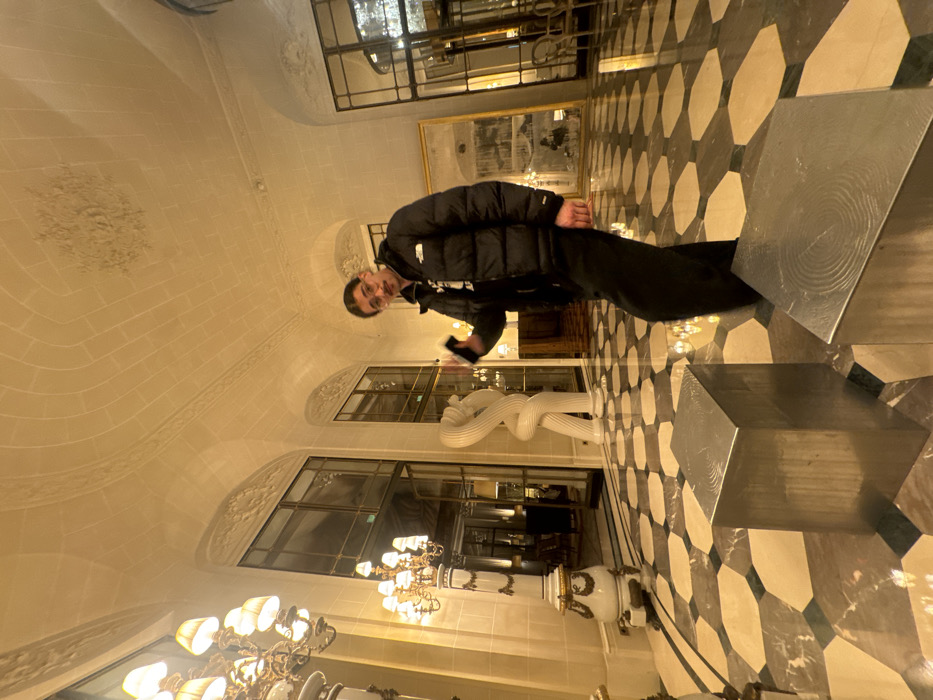 A young adult male stands centrally within the opulent lobby of the Hôtel de Crillon in Paris, France. He faces the viewer with a neutral expression, his dark hair swept back, featuring a thin mustache and goatee. He wears a black puffer jacket with a visible "The North Face" logo, a dark graphic t-shirt, and dark trousers. His left hand is raised slightly, holding a dark-colored smartphone.
The interior features a high, vaulted ceiling with intricate moldings and recessed panels, illuminated by warm artificial light. Large, arched windows or doorways with multiple panes line the walls, revealing softly lit interior spaces beyond. On the far left, a tall, ornate columnar stand supports a multi-branched chandelier. A distinctive white, twisted modern sculpture is positioned between two windows further left. To the right, a large, rectangular gold-framed artwork or mirror with a distressed, abstract surface leans against the wall. The floor is composed of highly polished, reflective marble tiles arranged in a geometric pattern of black, white, and grey octagonal and square shapes. In the immediate foreground, two rectangular, metallic silver-colored blocks, likely decorative elements, rest on the floor, their top surfaces exhibiting subtle concentric circular textures.