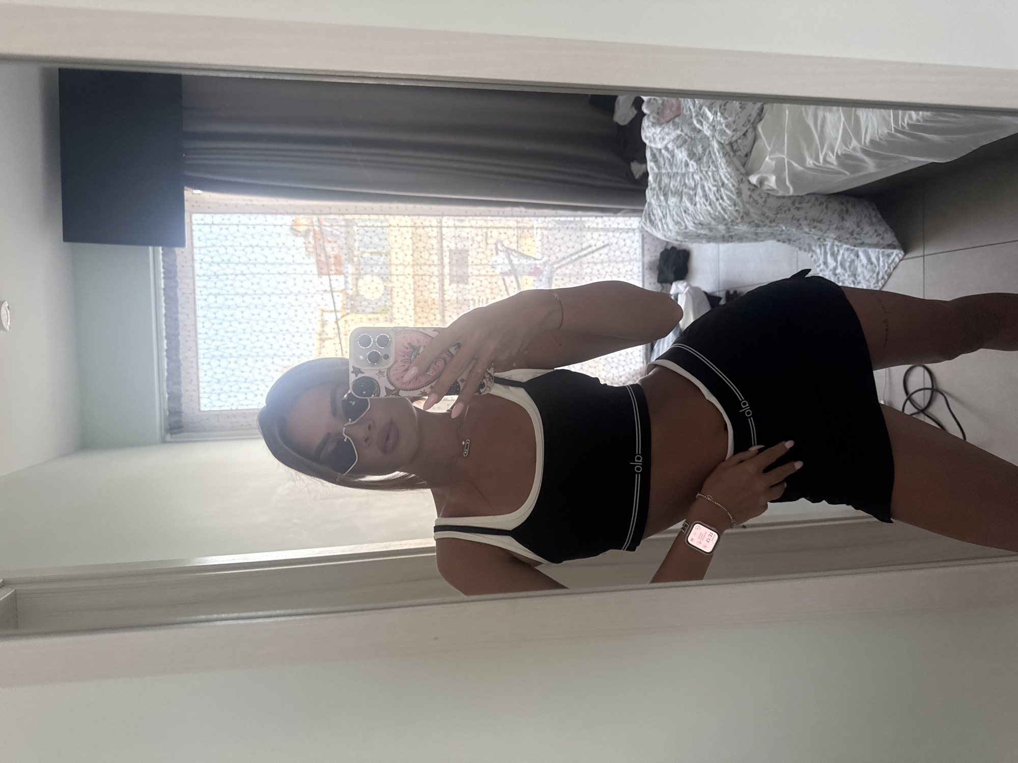 A young woman stands in a room, taking a mirror selfie. She is wearing a black sports bra with white trim and a matching black mini skirt. She has on heart-shaped sunglasses and a necklace. Her phone, adorned with stickers, is visible in her hand. The room appears to be a hotel room, with a made bed and a window with curtains. The overall impression is one of casual relaxation. The image was captured in Katerini Municipality, Greece.