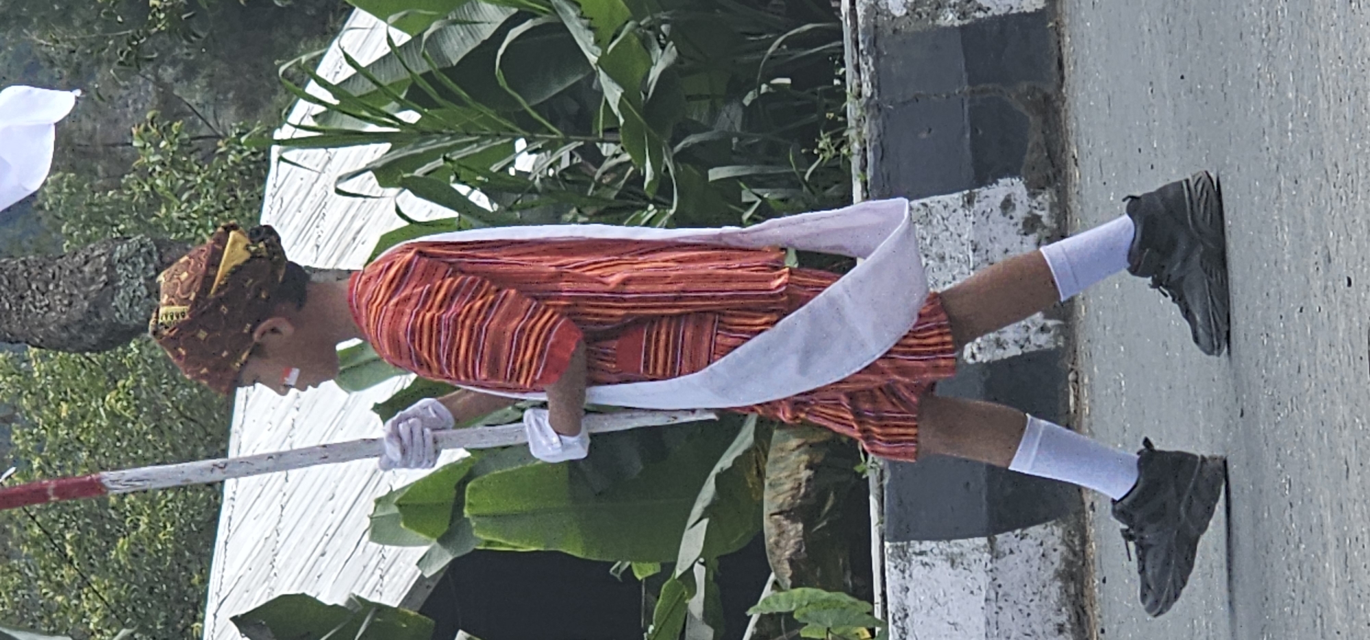 A young person, appearing to be a boy, is walking on a paved road, possibly as part of a parade or ceremony, in Puncak Jaya, Indonesia. He is wearing a traditional patterned head covering, an orange and red striped tunic and shorts, white gloves, white socks, and dark athletic shoes. A white sash is draped over his left shoulder and across his chest. He holds a white pole with red markings at the top in his gloved hands. His face has red and white face paint. In the background, lush green foliage, including large-leafed plants and trees, is visible. A white structure, likely a roof, is partially visible behind him. A concrete barrier with black and white stripes runs along the edge of the road. The lighting suggests it is daytime.