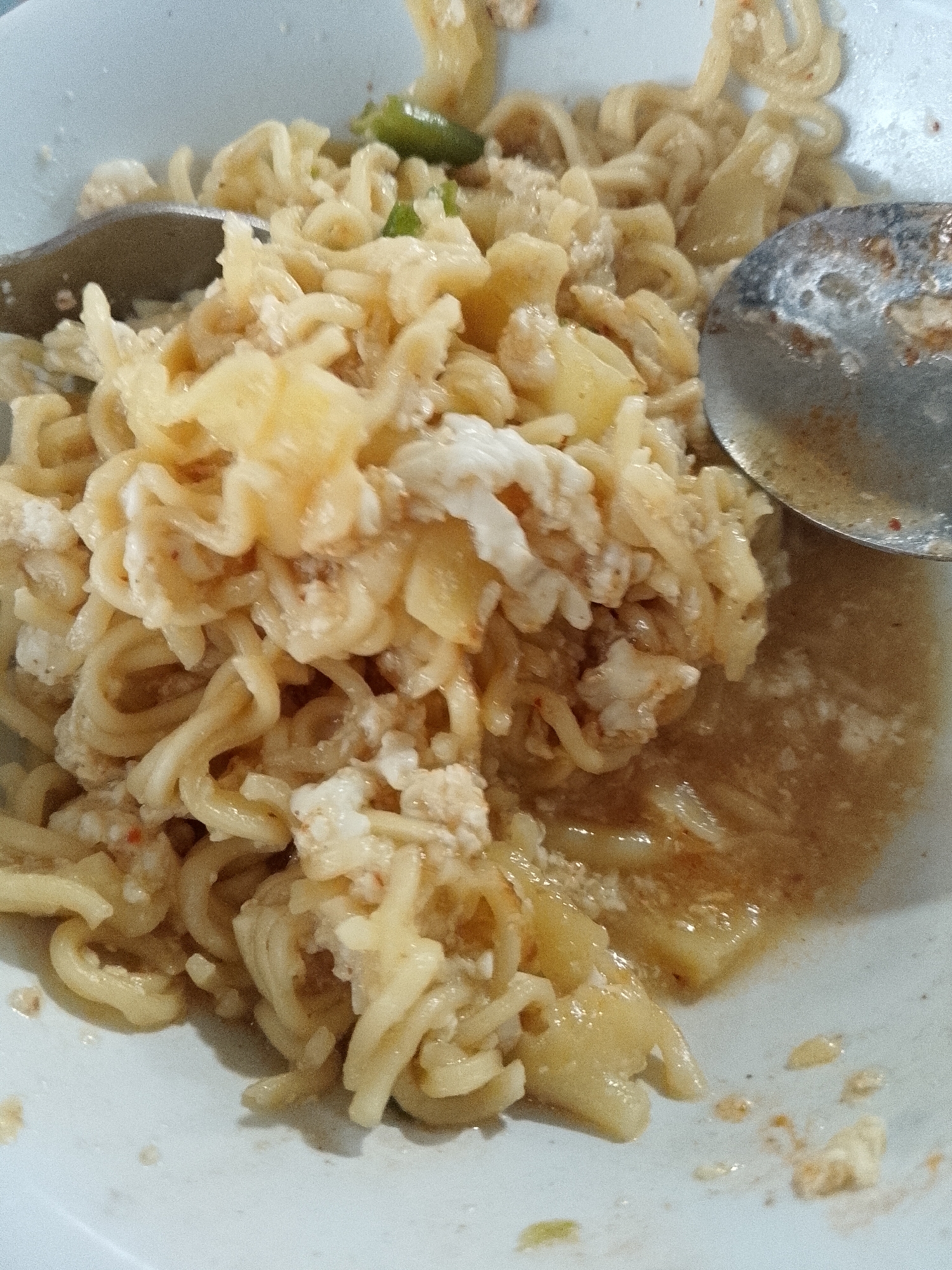 A close-up shot shows a plate of Mie Aceh, a noodle dish, likely taken in Lhokseumawe, Indonesia. The dish consists of thick yellow noodles mixed with what appears to be scrambled eggs, diced potatoes, and possibly a green chili. A light brown sauce covers part of the noodles, pooling at the bottom of the white plate. Two spoons, one containing remnants of the dish, rest on the plate amidst the noodles. The focus is on the texture and arrangement of the noodles and other ingredients.