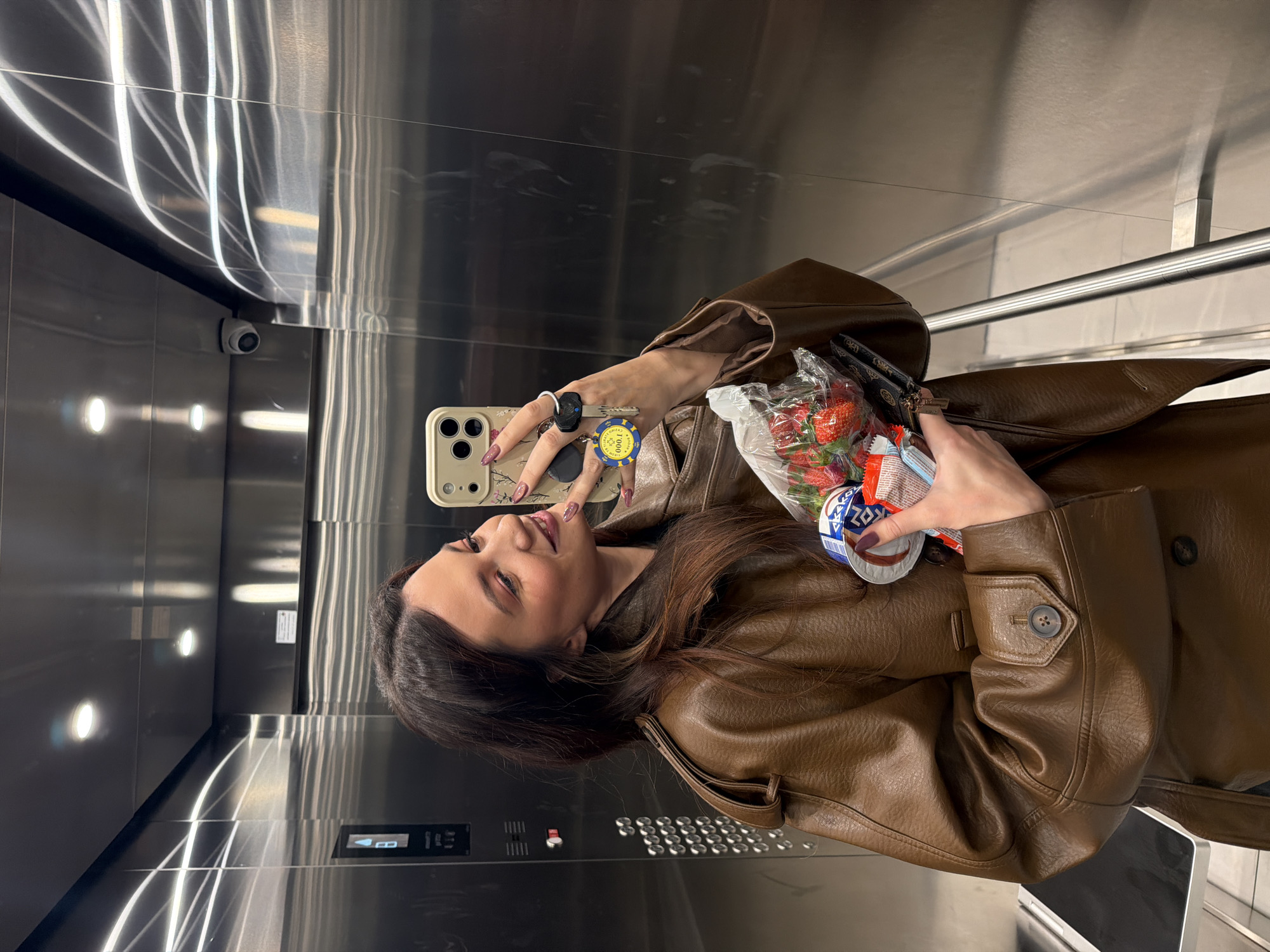 A woman in a brown leather jacket is taking a selfie in an elevator. She's holding a plastic bag containing strawberries and a container of sour cream. She is wearing rings on her fingers and has a phone with a beige floral case. The elevator has shiny metallic walls, a row of circular lights, and a metallic handrail. The image suggests the woman is in Belgrade, Serbia.