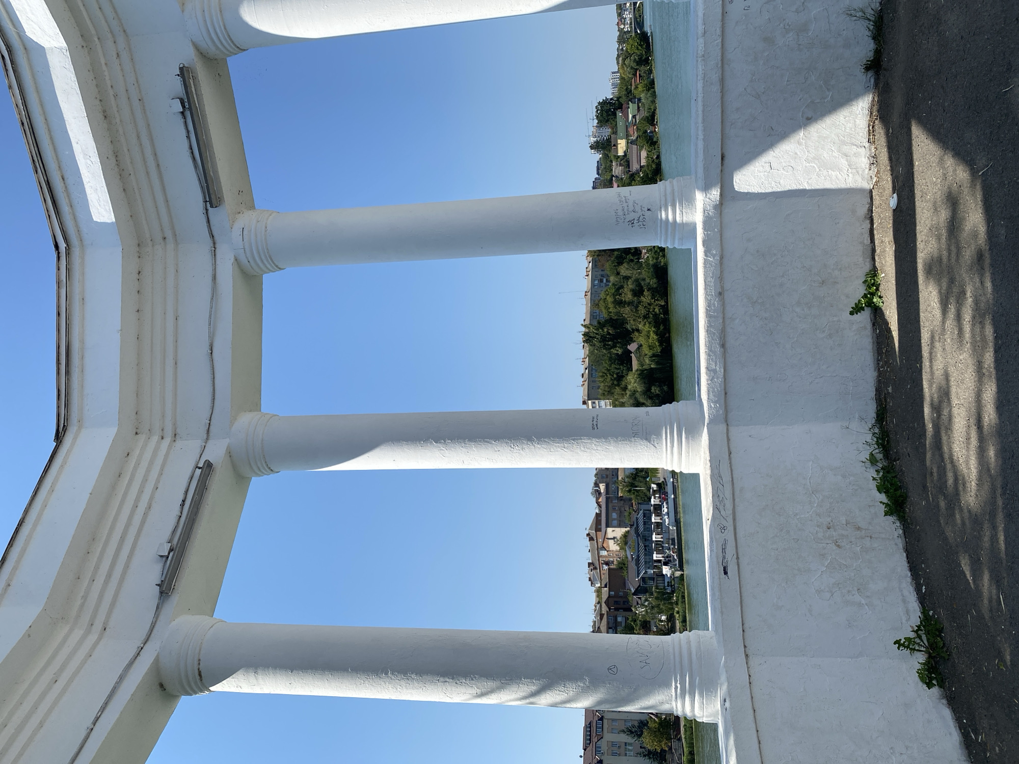 The image shows a view through a white, classical-style colonnade looking out over a river and city. The colonnade has at least five large white columns and a white balustrade. Some graffiti is visible on the columns and balustrade. In the background, across the river, is a cityscape with buildings, trees, and a clear blue sky. A boat is visible on the river. This scene is characteristic of the Vinnytsia National Technical University embankment in Vinnytsia, Ukraine, a popular recreational area that features architectural elements like colonnades overlooking the Southern Bug River. There are no people visible in the image.