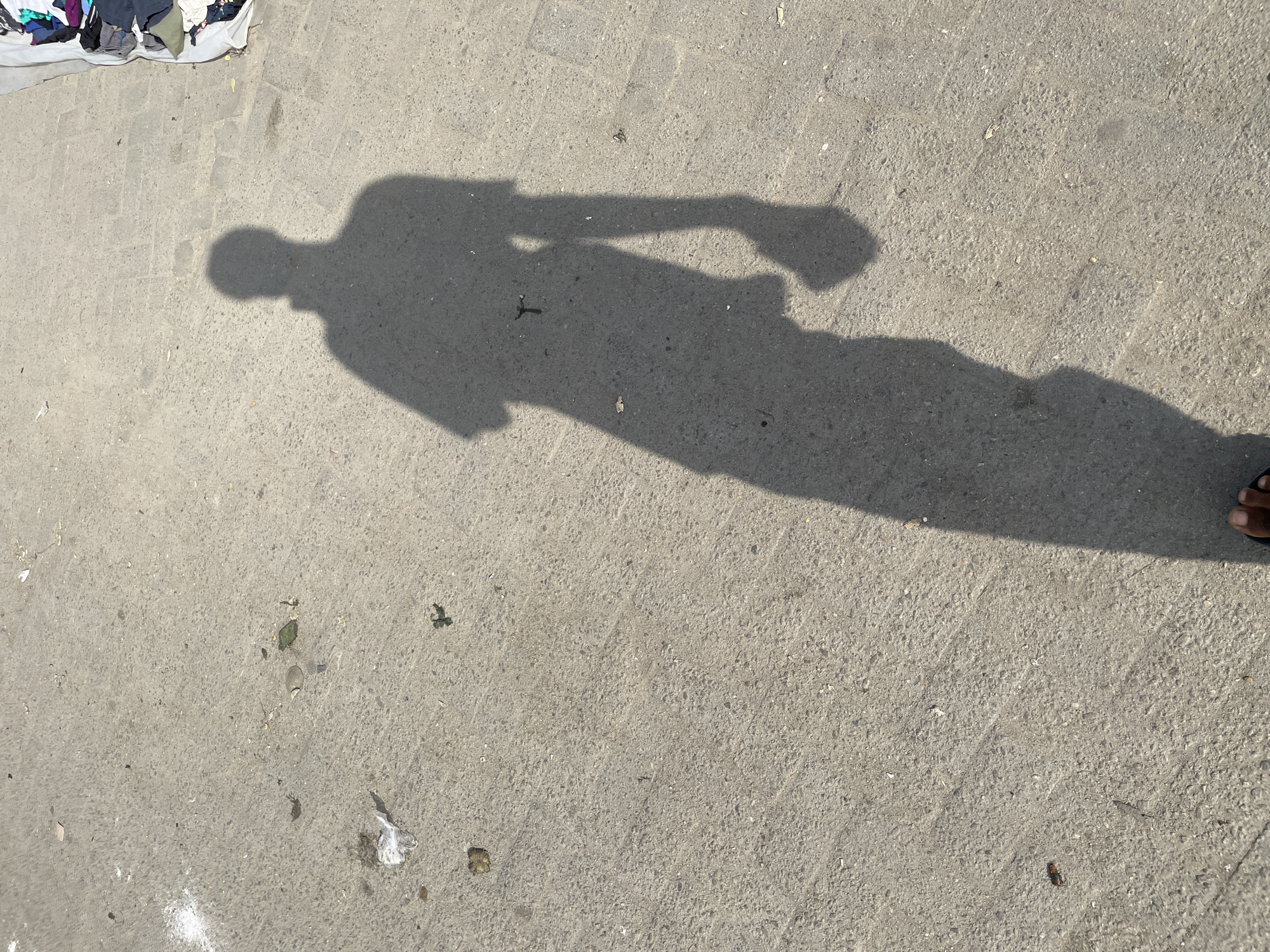 The image shows a shadow of a person standing on a paved ground. The ground appears to be made of concrete or asphalt and is somewhat rough and textured. There are scattered pieces of debris on the ground, including some white powder, plastic wrappers, and small dark specks. To the right of the shadow, a pile of folded clothes is visible, resting on a light-colored fabric. The shadow itself is long and distinct, indicating a strong light source, likely the sun, from the left. The figure in the shadow appears to be wearing a shirt and pants, with one arm bent and holding an object. The bottom of the image shows a portion of a person's feet wearing dark sandals. The scene is likely outdoors, given the shadow and the sunlight. The location is specified as a city in Lekki, Nigeria.