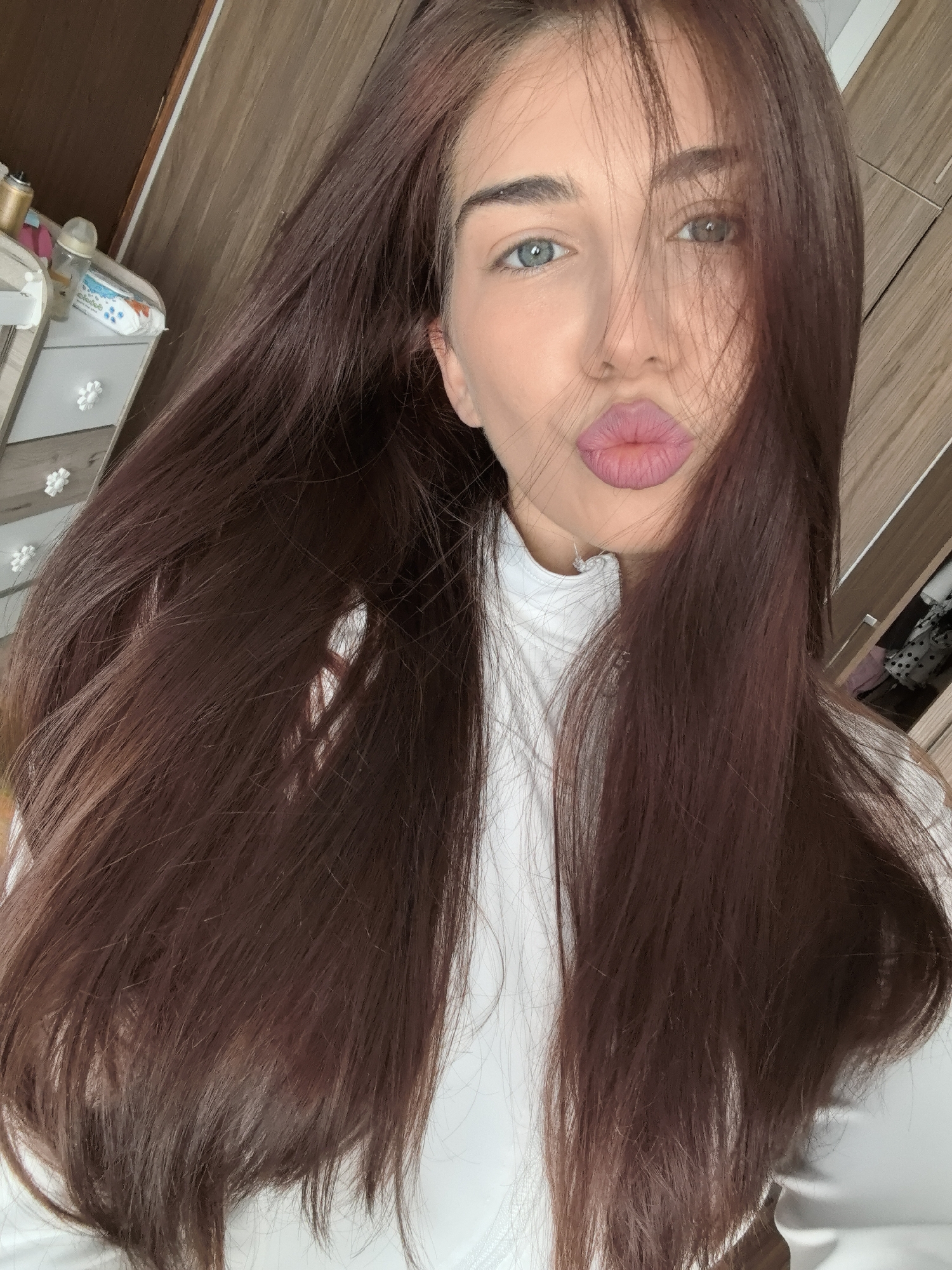 A young woman is taking a selfie. She has long, straight, auburn-colored hair that is partially obscuring her face. Her eyes are blue and she is pouting her lips which are painted pink. She is wearing a white turtleneck top. The background includes wooden furniture and what appears to be a changing table with baby supplies. The location cannot be determined to be Stara Zagora, Bulgaria, as there are no discernable clues.