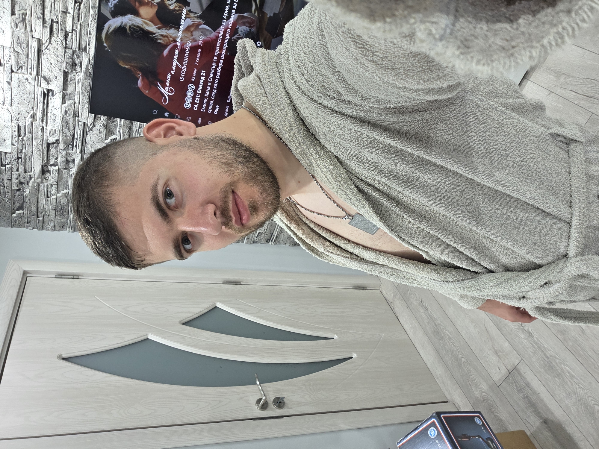 A man in a gray bathrobe stands indoors in front of a white door with decorative inserts. He has short hair and a beard. The man wears a silver necklace with a pendant. Behind him is a television showing what appears to be a program in Bulgarian called "Малки сладки лъжкини", or "Pretty Little Liars" in English. The wall behind the television is decorated with stone paneling. The floor appears to be light-colored wood. The man's exact location in the city of Yagodovo, Bulgaria, cannot be determined from the image.
