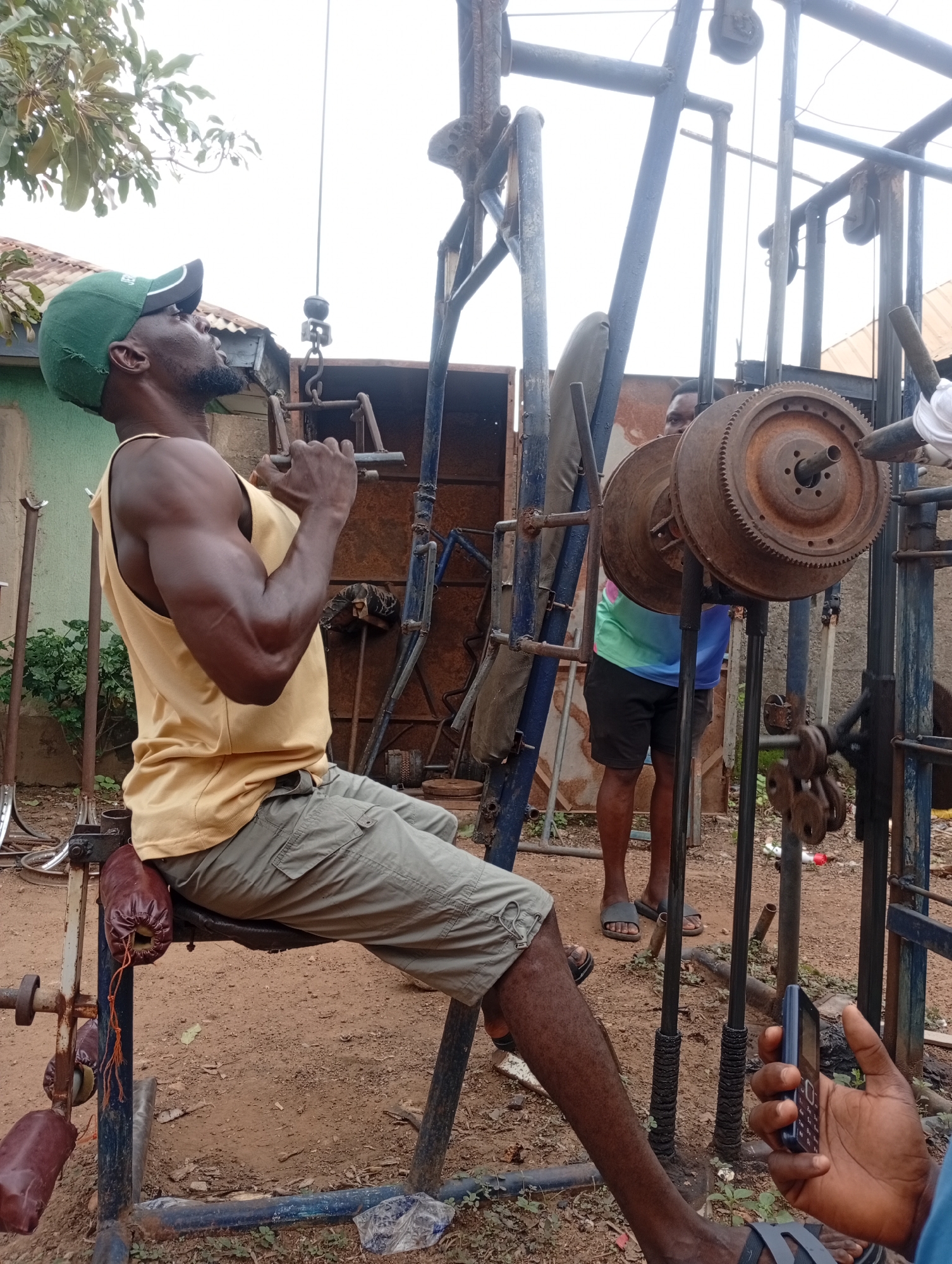 This image captures an outdoor fitness scene in Akwanga, Nigeria, centered around a homemade gym setup. The primary subject is a dark-skinned man, seen in profile from the left, engaged in an exercise that appears to be a lat pulldown or row. He is seated on a crude, padded bench, wearing a green baseball cap, a yellow tank top that reveals his muscular arms, and khaki cargo shorts. His eyes are cast upwards, reflecting concentration on his workout.
The exercise equipment is distinctly homemade, constructed from robust, possibly recycled, metal pipes, chains, and large, rusty industrial gears serving as weights. This DIY approach highlights resourcefulness. In the background, another dark-skinned man, dressed in a colorful short-sleeved top and black shorts, is partially visible, observing the workout. A third person's hand, holding an older model cell phone with a physical keypad, is seen in the bottom right corner of the frame.
The setting is an unpaved, outdoor area with reddish-brown earth, common in the region. Various metal parts and objects are scattered on the ground. Behind the equipment, rudimentary structures, possibly sheds or unfinished buildings with corrugated iron roofs and painted walls, are visible. Green trees and foliage are present, suggesting a natural environment surrounding the exercise area. The sky above is bright and appears slightly overcast, indicating daylight. The scene conveys a sense of determined effort and community in a rustic, functional fitness environment. No discernible text is visible on clothing or equipment.