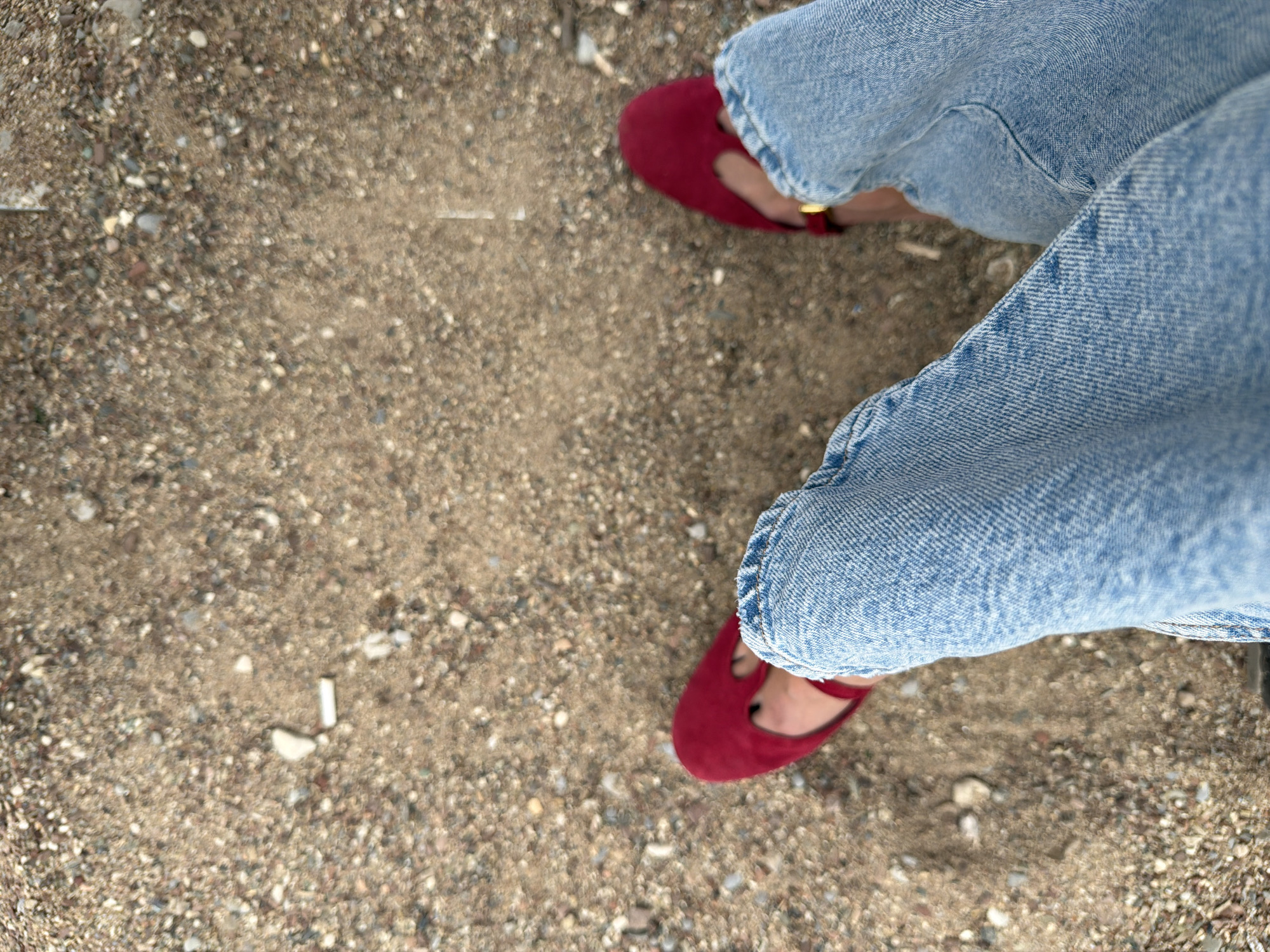 The image shows a view from above of a person's legs and feet standing on a gravelly or sandy surface. The person is wearing blue denim jeans with the cuffs rolled up and red suede Mary Jane-style shoes. The surface underfoot appears to be a mixture of sand, small pebbles, and some bits of debris. A cigarette butt can be seen on the surface. Given the ground texture and the possibility of a beach setting, this image may be from the city of Budva, Montenegro. More specific information about the location would need additional visual cues.