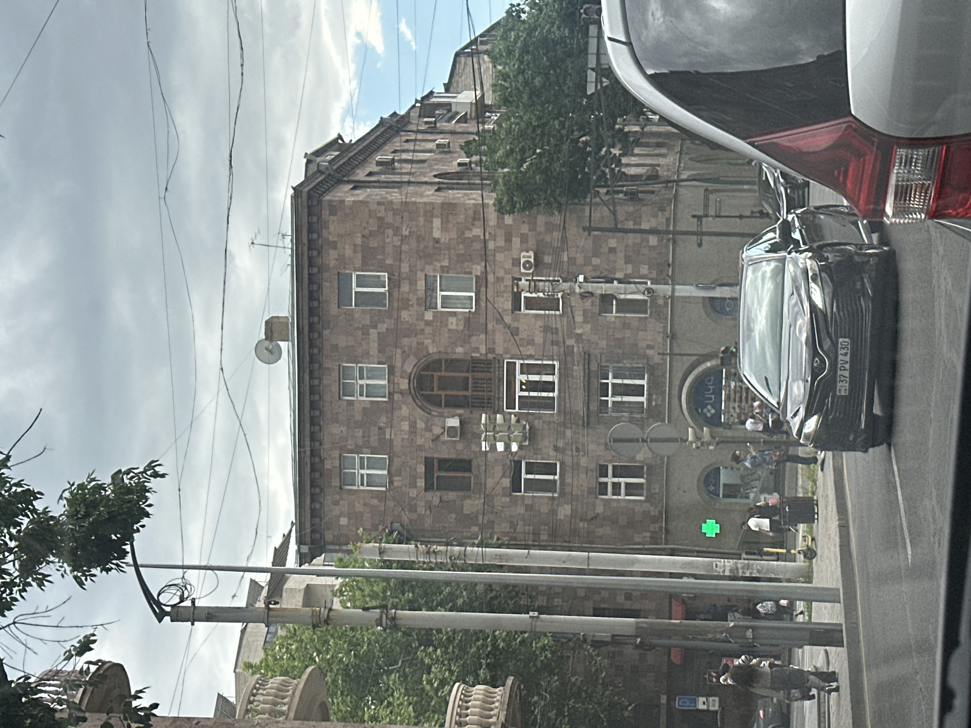 The image is taken from inside a vehicle, showing a street scene in Yerevan, Armenia. A multi-story building with a facade of brown stone blocks dominates the view, featuring multiple windows, including a distinctive arched window. A black car is partially visible, its license plate reading "37 PV 430". Street signs, utility poles, and power lines are prominent. There are people on the sidewalk near the building, and a green cross is visible. The sky is overcast with clouds.