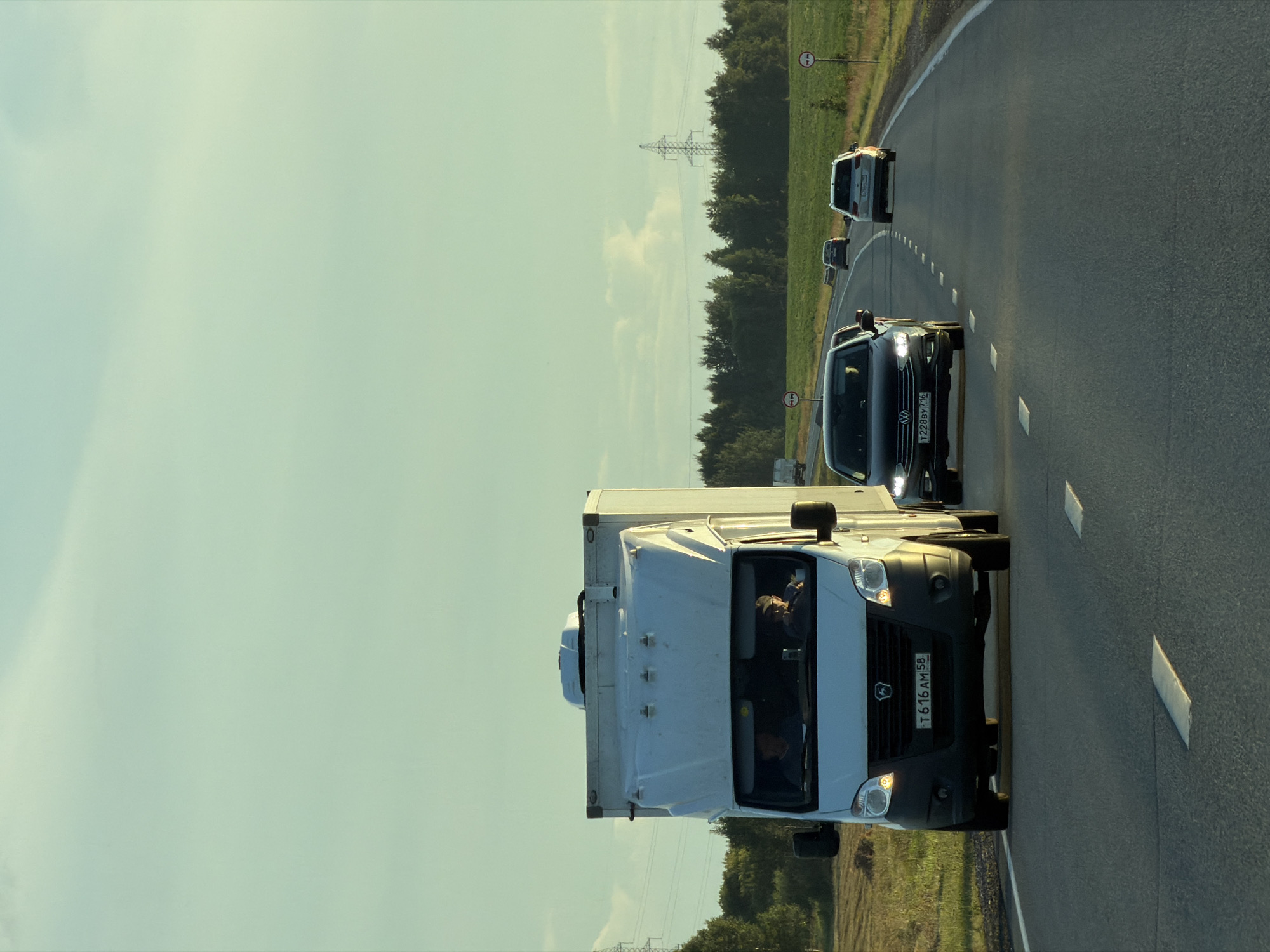 A white truck with a box trailer is traveling on a highway. Inside the truck, two men are visible in the cab. The truck is in the left lane of a two-lane road. To the right of the truck, a black sedan is traveling in the same direction. Further ahead, a white SUV is also on the highway. The road is paved with asphalt and has white lane markings. On the right side of the road, there are fields of grass and trees. Power lines and a transmission tower are visible in the background. The sky is clear with a pale blue hue. The scene appears to be set in a rural or semi-rural area, possibly Togashevo, Russia, given the context.