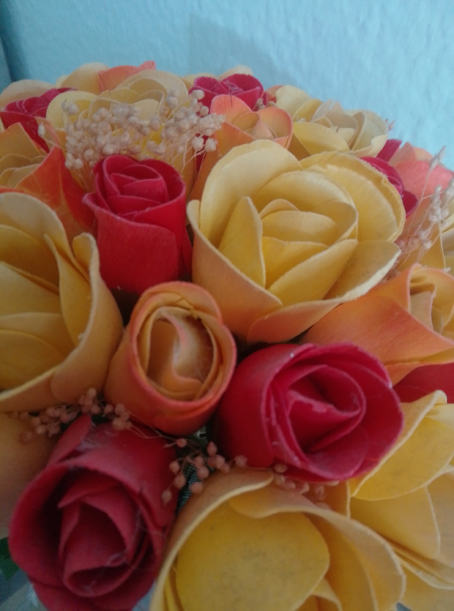 A bouquet of artificial roses is displayed against a light blue background. The roses are a mix of yellow and red, with some lighter orange-pink hues. Small, pale sprigs are interspersed between the roses. The bouquet appears to be a single object, with no people or further context provided. There is no information in the image that allows for the determination of its location in Wedel, Germany.