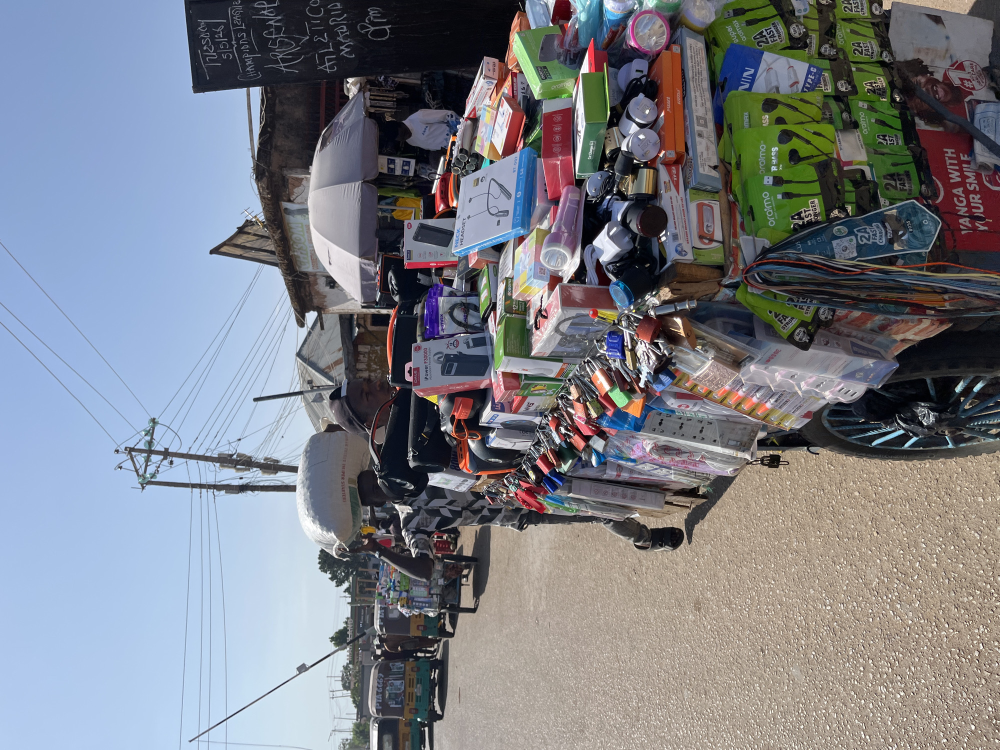 A street vendor in Gura Top, Nigeria, walks along a paved road, carrying a load of goods, including flashlights, phone chargers, and headphones, displayed on a wheeled cart. The vendor wears a black and white patterned shirt and has a large sack on their head. Motor tricycles are parked nearby. Overhead, utility poles and power lines extend along the street. In the background, building with a sign reading "Tuesday 5/5/26" and "ARSENAL" are visible.