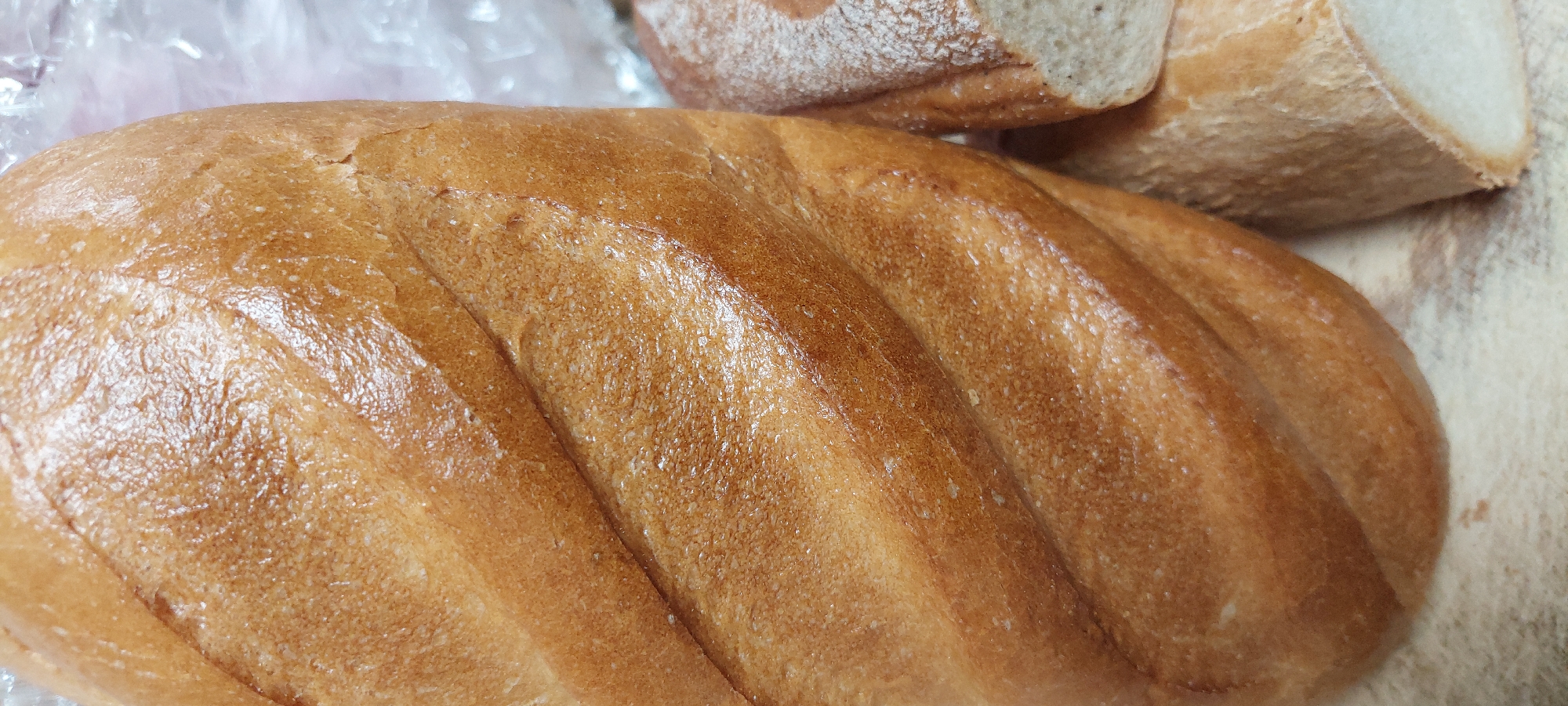 A close-up, eye-level, vertical shot shows two loaves of bread. The loaf on the left is a light brown, whole loaf with distinct scoring marks running diagonally across its surface. The loaf on the right is partially visible, showing its end slice, which is a lighter, cream color with a dusted topping of flour. The bread appears to be fresh, with a slightly glossy crust on the left loaf. The background is blurred and appears to be a light-colored surface, possibly a countertop, with some indistinct white material to the right of the loaves, which could be plastic packaging. There are no people or other identifying features in the image. The location is unknown.