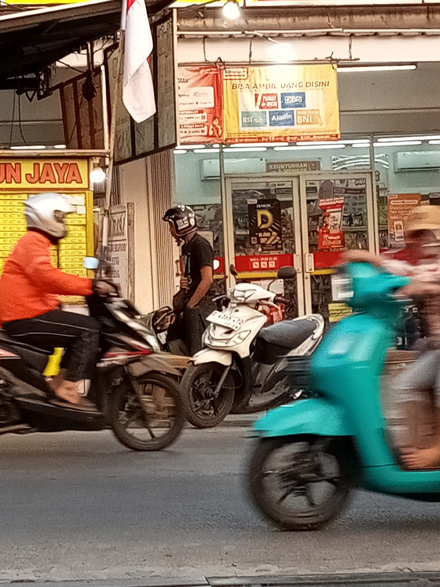 This is a street-level, medium shot photograph taken outdoors in Sariwangi, Indonesia. The scene depicts a busy commercial area with several individuals on motorcycles. In the foreground, a person wearing an orange jacket and a white helmet is riding a motorcycle. To their right, another person in a black shirt and helmet is standing next to a white motorcycle. A turquoise scooter is blurred in motion on the right side of the frame. Behind the individuals and motorcycles, a storefront is visible, featuring signs with Indonesian text and logos of banks, indicating a financial service point. The overall atmosphere suggests a typical urban environment with active street life.