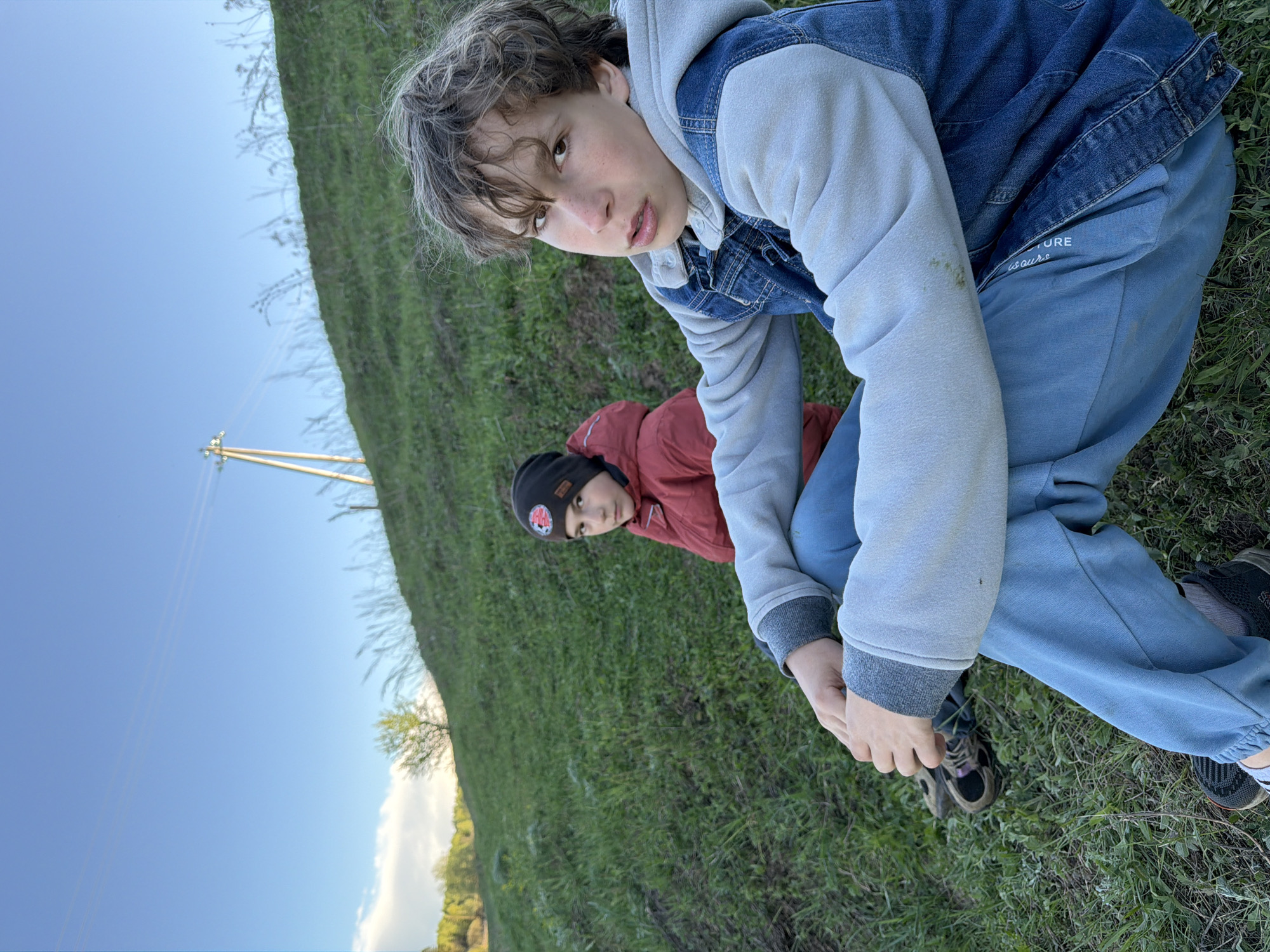 Two young boys are outdoors in Фоки, Russia. The main subject, a boy with curly hair, is positioned in the foreground, looking at the camera, wearing a grey hoodie, blue denim jacket, and blue pants. He is crouched down on grass. Behind him and slightly to the left is a second boy wearing a red jacket and a black beanie. The ground is covered in grass. In the background, there is a clear blue sky and a utility pole with power lines is visible. There is also some vegetation including trees.