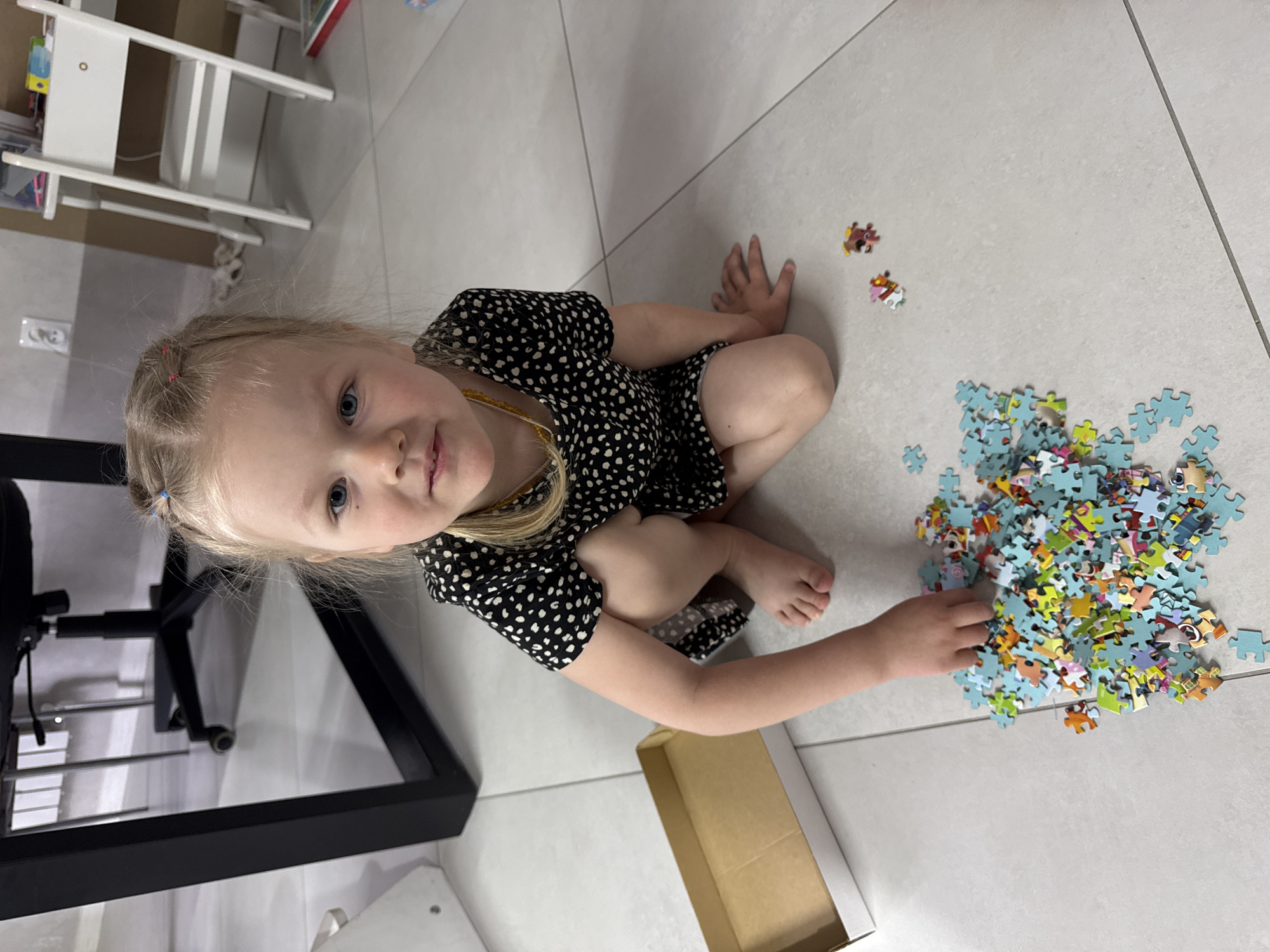 A young blonde girl is captured from an overhead perspective, sitting on a light-colored tiled floor indoors, likely in Gornyi (Dvurechenskoe S/P), Russia. She is wearing a black dress with white polka dots and a yellow beaded necklace. Her blonde hair is styled in two small pigtails, each secured with a pink elastic band, and her blue eyes look directly at the viewer with a calm, neutral expression.
The girl is engaged in an activity with a large pile of colorful puzzle pieces scattered on the floor to her right. Her right hand rests among the pieces, which display various images and have blue backings. A few individual puzzle pieces are slightly separated from the main cluster. An empty, light brown cardboard puzzle box lies on the floor near her hand.
In the background, a white multi-tiered shelf or storage unit is visible behind her to the upper left. A black office chair with wheels is partially visible to her left. The floor consists of large, light grey or white tiles with visible grout lines, suggesting a modern indoor setting. The clear lighting indicates it is daytime. No discernible text is visible in the image. The scene depicts a quiet moment of play and concentration.