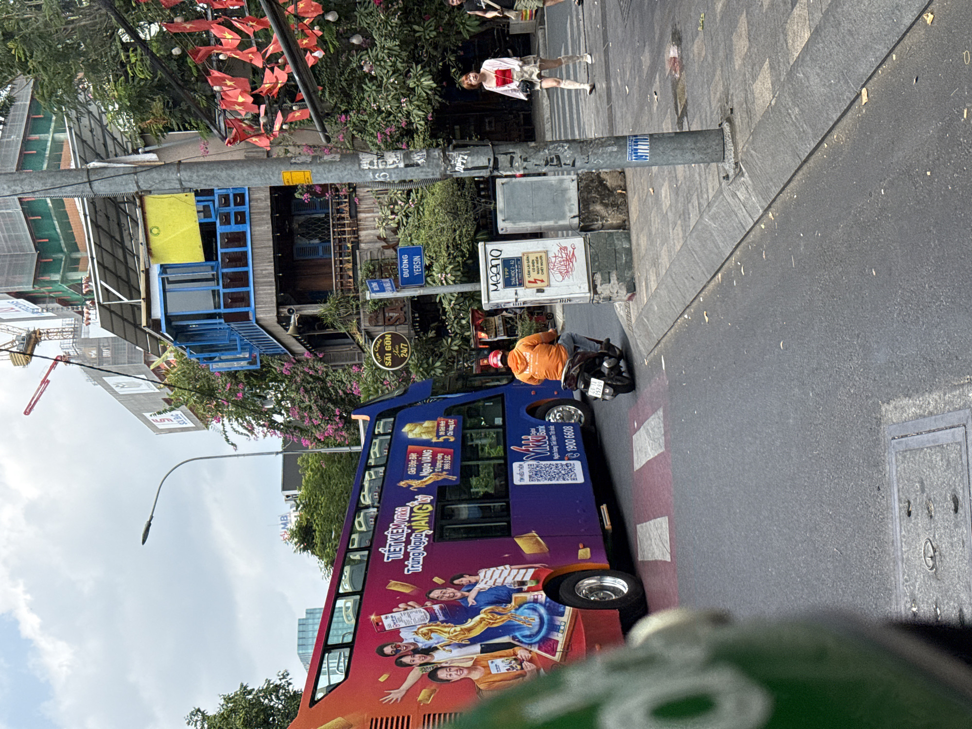 An eye-level shot captures a vibrant street scene in Thủ Đức, Vietnam, during daytime under an overcast sky. The image is oriented vertically, looking along the street.
In the foreground, a colorful double-decker bus dominates the lower frame, adorned with large advertisements. The bus is painted purple and orange, featuring images of people joyfully interacting with gold bars and a golden horse. Visible text on the bus includes "MB Bank," "TIẾT KIỆM VÀNG" (Saving Gold), "Giải Đặc Biệt Trúng Ngựa Vàng" (Special Prize Winning Gold Horse), and a phone number "1900 6608."
Behind the bus, a person in an orange jacket is seen on a motorbike. To the right, a wide sidewalk features a woman standing, dressed in a white top with a red undershirt and light-colored shorts. Another person is partially visible behind her. The sidewalk is separated from the asphalt road by a concrete curb.
Numerous street poles are visible, bearing signs and wires. One blue sign clearly reads "ĐƯỜNG YERSIN," identifying the street. Below it is another sign for "SAI GON Cà phê 24/7." A utility box nearby has graffiti and text such as "MEENO," "TPP," "THÁI HỌC 2-A2," and a striking phrase "CẨM LẠI GẦN CHẾT NGƯỜI" (Stay close to kill people). Overhead, a dense canopy of small red flags with yellow stars (Vietnamese flags) is strung across the street, adding a festive or patriotic touch. Lush green trees and flowering plants line the street, bordering various buildings, some with blue-framed windows. A construction crane is partially visible in the distance, indicating ongoing development.