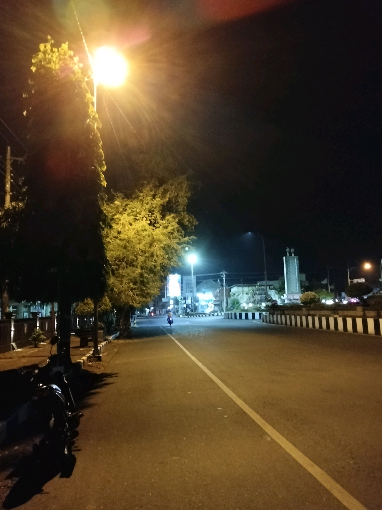The image depicts a street scene at night in Purworejo, Indonesia. A bright street lamp illuminates a tree on the left side of the road, casting long shadows. The road itself is paved and has a white line marking the lane. On the left side of the road, there are parked motorcycles and a low wall with decorative elements. In the distance, on the right side of the road, there is a monument with statues on top, a roundabout with black and white striped borders, and several buildings. A single person is seen riding a motorcycle down the street. The overall atmosphere is quiet and dimly lit, characteristic of a nighttime urban environment.
