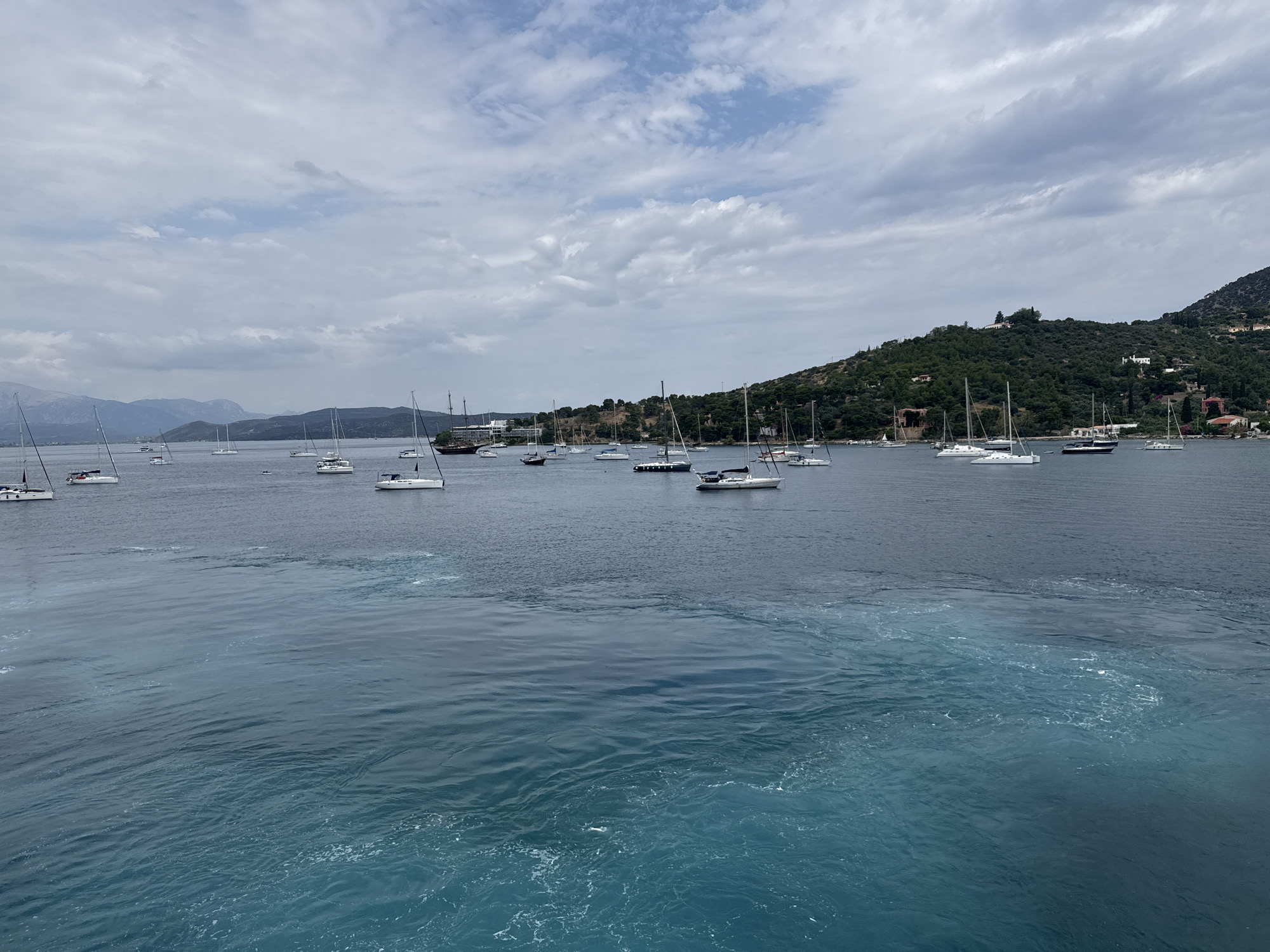 The image shows a bay in Poros, Greece, filled with numerous sailboats and yachts. The water in the foreground is a vibrant turquoise, transitioning to a deeper blue further out. The bay is surrounded by a green, hilly coastline with scattered buildings and vegetation. In the distance, mountains are visible on the horizon. The sky is partly cloudy with a mix of white and gray clouds.