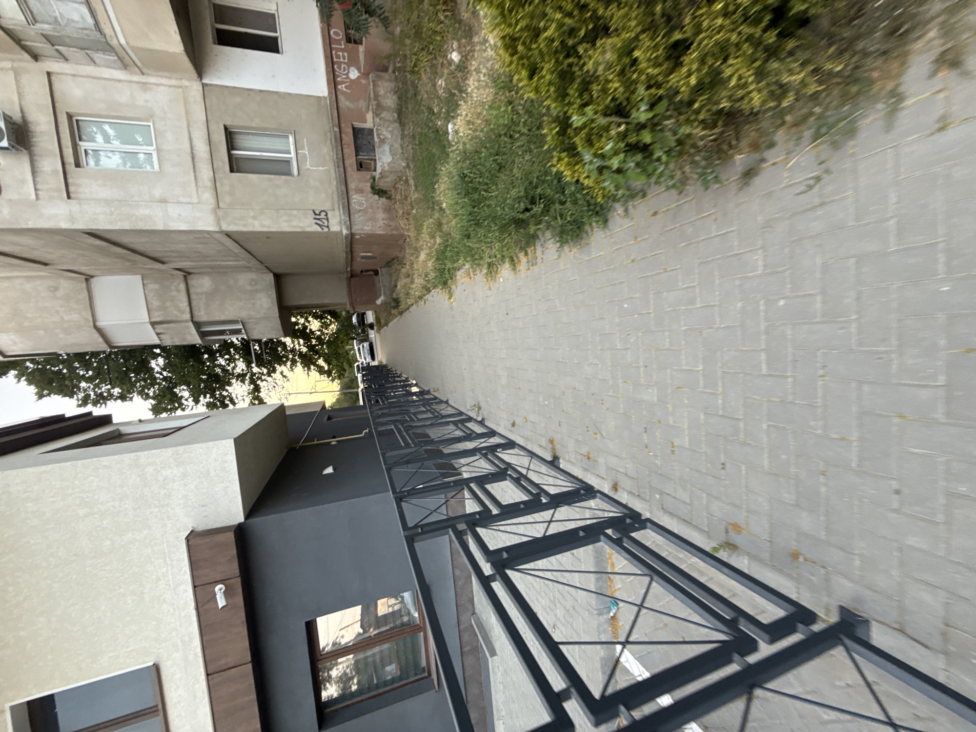 This media file captures an outdoor scene in Cahul, Moldova. The image is oriented vertically, with the foreground featuring a modern building constructed with light-colored and dark gray textured facades. This building has several windows and architectural details like dark brown trim. A black metal fence with a geometric design runs along the side of the building, bordering a paved walkway. The walkway is made of gray rectangular bricks laid in a herringbone pattern.
Beyond the walkway, there is a patch of overgrown green and dry grass, with some scattered debris. Further in the background, an older, multi-story building is visible. This older building has a rough, beige-colored exterior and features several windows, some with white frames and others with darker frames. Graffiti is visible on the exterior of this older building, including the word "ANGELO" written vertically in black, and the numbers "115" and "STF" also appear to be inscribed on the wall.
To the left of the older building, trees with green foliage are present, suggesting it is daytime and likely a temperate season. The lighting suggests it is either morning or late afternoon, with soft shadows. The overall atmosphere is quiet and urban, with a juxtaposition of newer and older architecture. There are no people visible in the frame, and the focus is on the architectural elements and the surrounding environment. The scene appears to be a residential or mixed-use area.