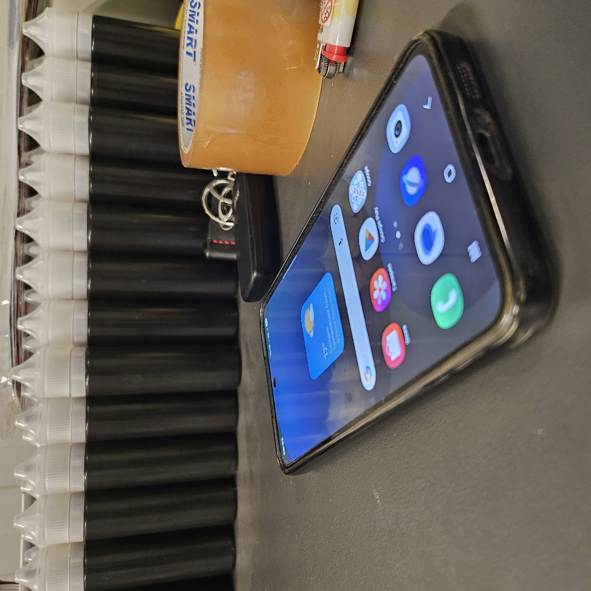 A smartphone lies on a dark, textured surface in the foreground. The phone's screen displays a blue background with weather information, indicating 17 degrees and a cloud icon, along with icons for various apps including "Store," "Gallery," and "Google Play." A search bar is visible at the bottom of the screen.
Behind the phone, a roll of clear packing tape is positioned to the right. To the left of the tape and further back, a black key fob with a metallic keychain is visible. In the background, a row of identical black cylindrical bottles with white, pointed caps are lined up.
The environment appears to be indoors, with the objects suggesting a storage or workspace. There is no indication of the specific location within Nowa Wieś Książęca, Poland. The scene is static and there are no individuals present.