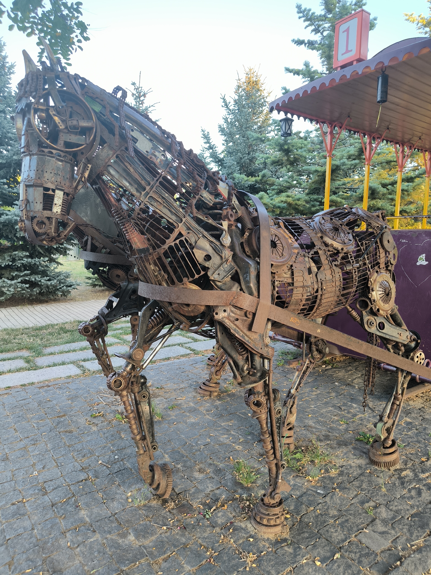 A life-sized sculpture of a horse, constructed from various scrap metal parts and gears, stands in an outdoor setting. The sculpture is detailed, with visible springs, chains, and interlocking metal components forming its body, legs, and head. The horse is positioned on a paved area with interlocking stone tiles, interspersed with patches of grass and fallen leaves.
Behind the sculpture, a portion of a covered structure, possibly a gazebo or a waiting area, is visible. This structure has a maroon-colored roof with a scalloped edge, supported by yellow posts. A lantern hangs from the underside of the roof, and a red sign with the number "1" in a white square is prominently displayed. A dark purple wall or barrier is partially visible to the right of the horse sculpture, bearing some faint graffiti or markings.
The background consists of lush green evergreen trees, suggesting a park or garden environment. The sky is a pale blue with a slight haze, indicating it may be late afternoon or early morning. The overall impression is one of a public art installation in a recreational area. The location is identified as a park in Chișinău, Moldova, known for its artistic metal sculptures.