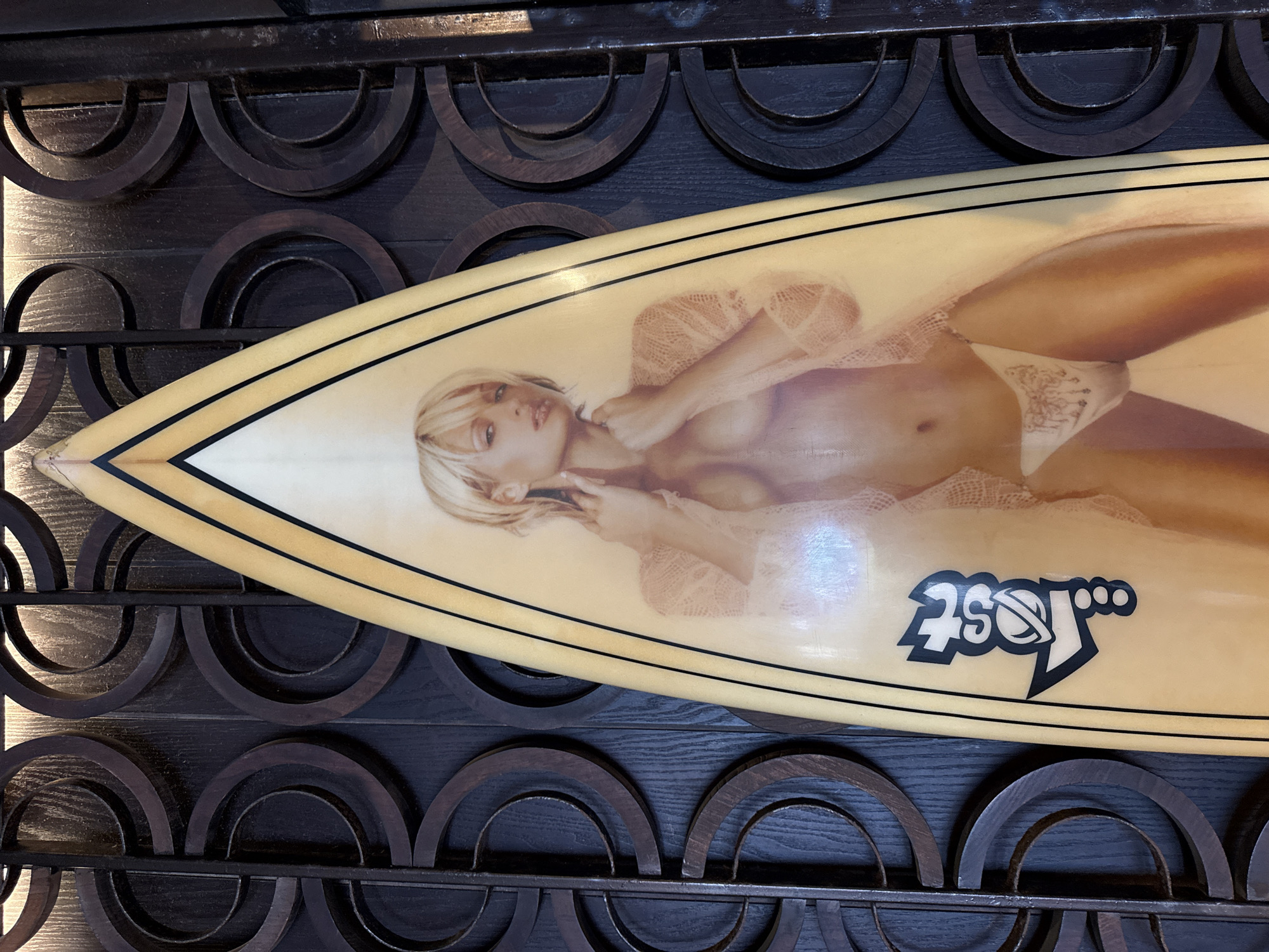 A cream-colored surfboard with black pinstriping and a faded image of a blonde woman wearing lingerie is displayed against a dark brown wooden decorative wall panel featuring repeating semi-circular designs. The surfboard has a pointed nose and a black "Lost" logo near the tail. The image appears to be taken in a retail or display setting. The specific location within Kuta, Indonesia, is not discernible from the image.