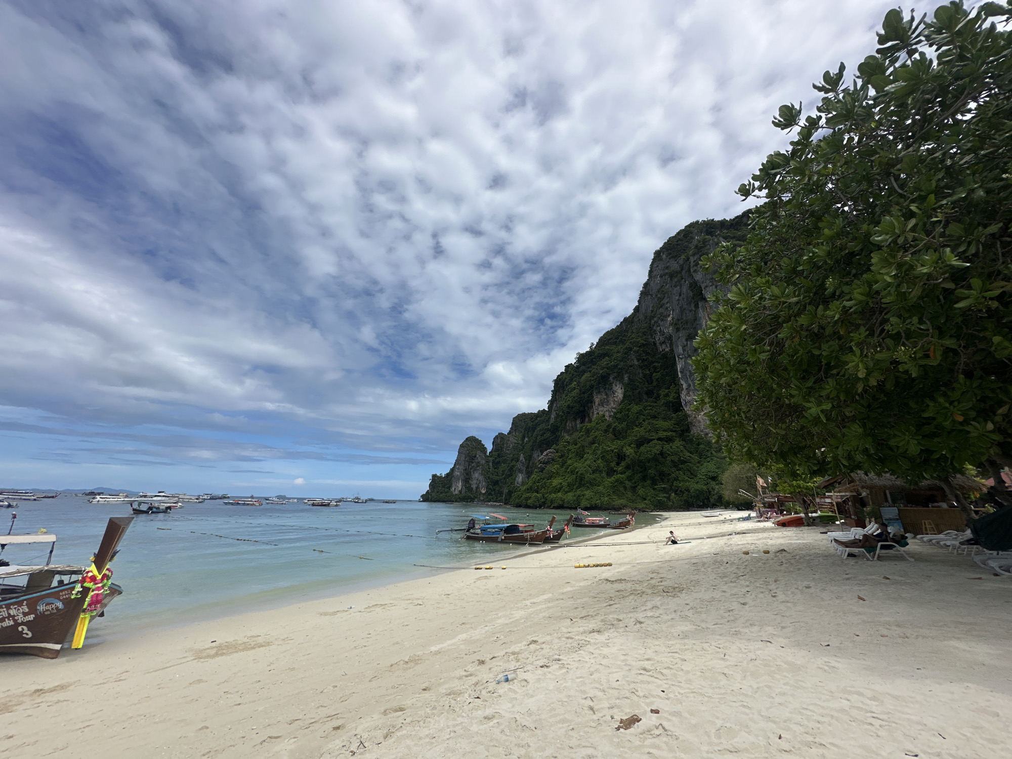 The image depicts a tropical beach scene in Ao Nang, Thailand, characterized by a white sand shoreline meeting a calm, clear turquoise sea. Dominating the background is a sheer, verdant limestone cliff, characteristic of the region. Numerous longtail boats, a local mode of transport, are moored in the shallow water, with some closer to shore and others further out. On the beach, there are several lounge chairs, some occupied by sunbathers, and a small thatched-roof bar or kiosk. A single person is visible sitting on the sand near the water's edge in the mid-ground, and another person is lying on a lounge chair in the foreground right. The sky is partly cloudy with wispy formations against a blue backdrop. The overall atmosphere suggests a leisurely day at a popular tourist destination.