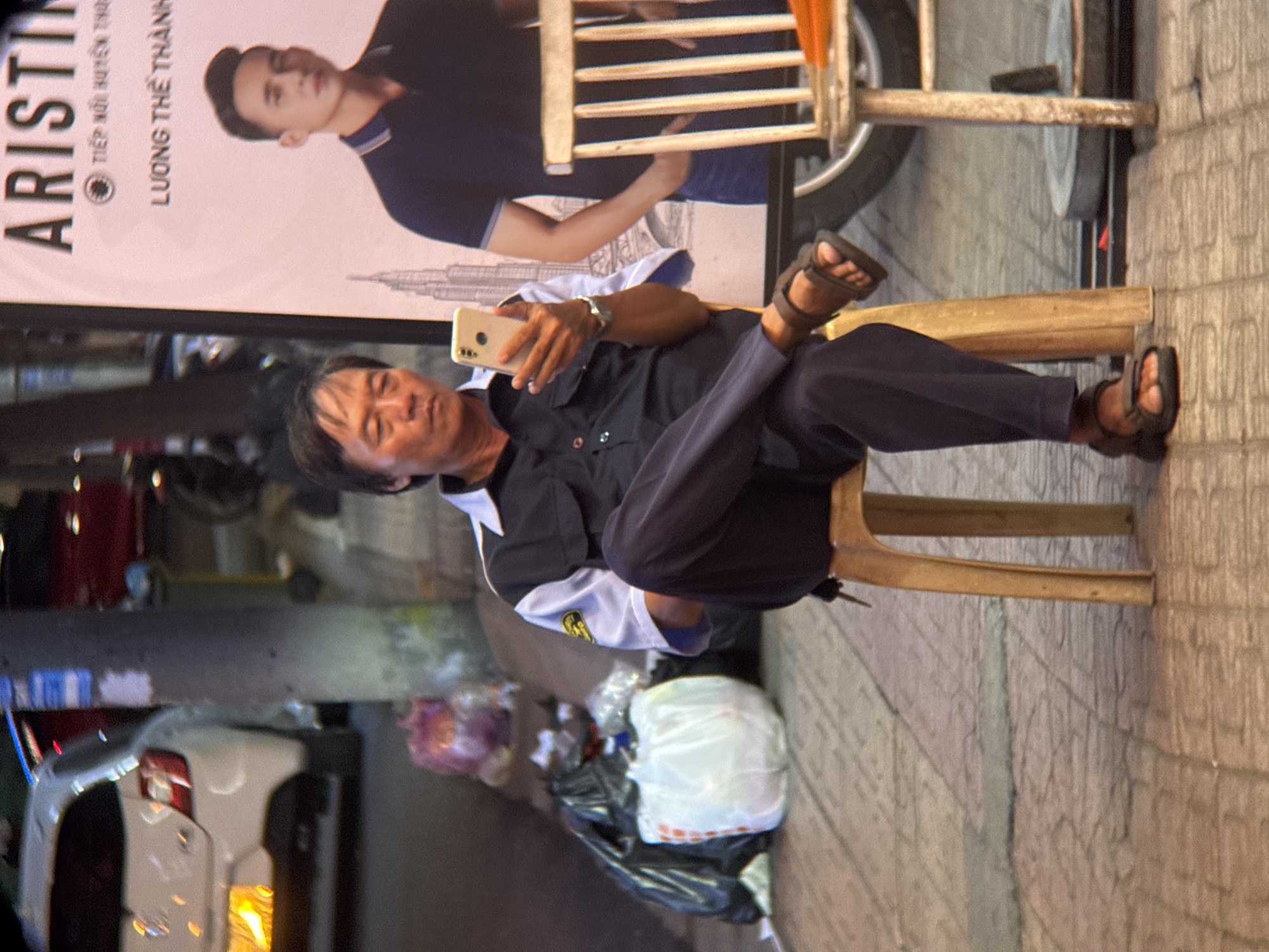 A man is sitting on a plastic chair outdoors, in what appears to be a street scene in Nha Trang, Vietnam. He is holding a smartphone in his right hand and looking at it. The man is wearing a dark blue short-sleeved shirt with white and yellow accents on the collar and sleeves, and dark blue pants. He is also wearing dark sandals. The background shows a white car, a lamppost, a large poster with text and an image of a man, and some trash bags. The ground is paved with patterned tiles. The overall impression is that of a casual moment captured on a street in a Vietnamese city.