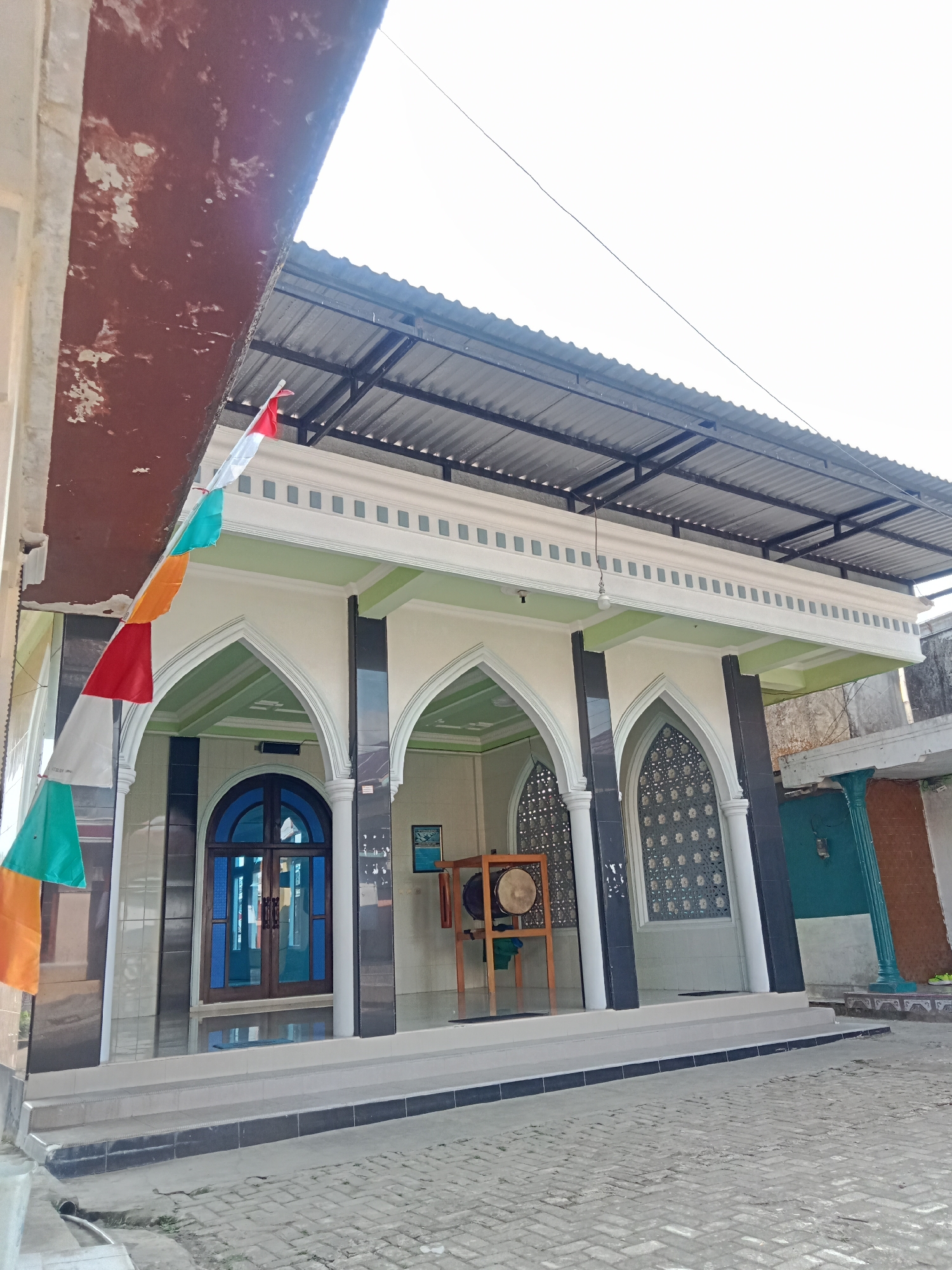 A wide shot shows the exterior of a building that appears to be a mosque in Pancurwening, Indonesia. The building has a white facade with arched entrances and decorative elements. The roof is corrugated metal supported by a dark frame. A string of colorful triangular flags, red, white, green, and orange, hangs in the foreground. In front of one of the arched entrances is a large, round drum on a wooden stand. The ground in front of the building is paved with light-colored bricks. The sky is bright and overcast.