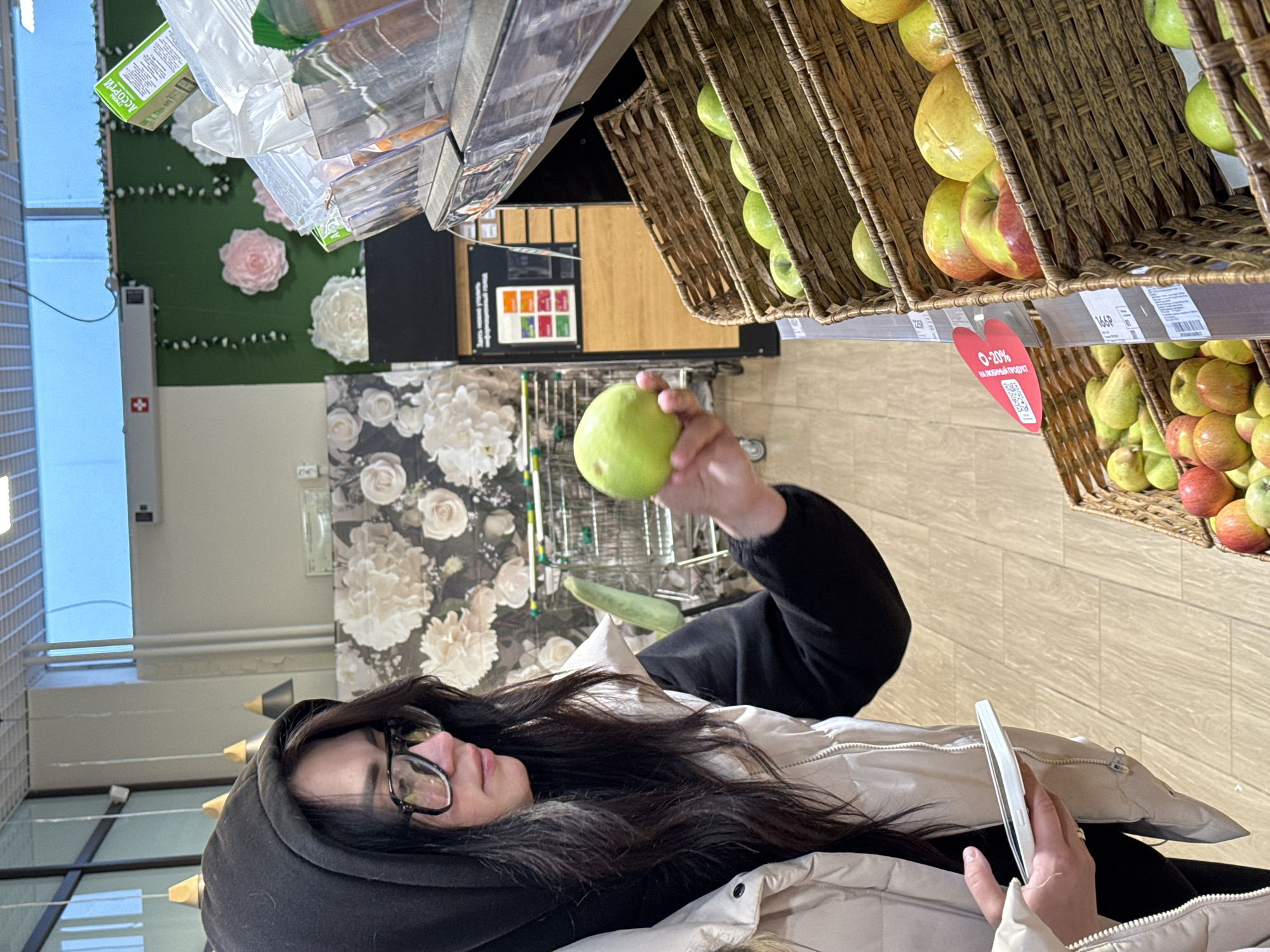 A woman is holding a green pear in a grocery store. She is wearing a black hoodie and a beige jacket. She has dark hair and glasses. She is holding a phone in her left hand. The store has baskets of apples and pears. The wall behind her has a floral design.